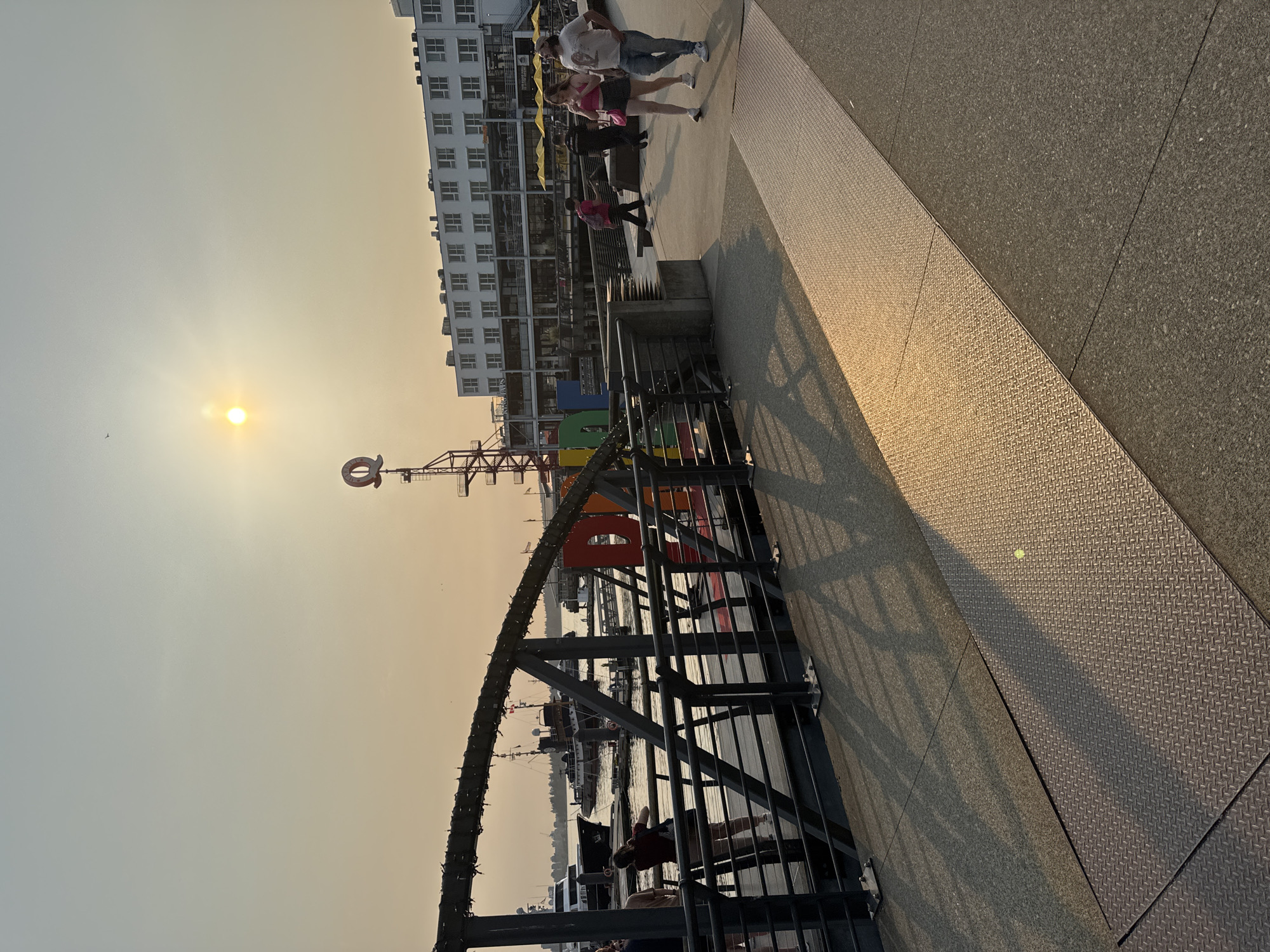 The image depicts a waterfront promenade in North Vancouver, Canada, bathed in the warm, hazy light of a setting sun. The sky is a pale orange and yellow, suggesting smoky conditions. In the foreground, a grey concrete walkway is partially covered by a diamond-patterned metal plate. A dark metal railing runs along the edge of the promenade, casting long shadows across the walkway. Beyond the railing, a body of water is visible, dotted with various boats and yachts, including a large black vessel and a more traditional-looking ship.
A colorful art installation, appearing to spell out a word with large block letters in shades of red, orange, yellow, green, and blue, is situated on the waterfront. Above this installation, a tall metal structure with a large red circular logo at its peak is visible.
Several people are present on the promenade. In the middle ground, a couple walks towards the right side of the frame, the woman in a pink tank top and skirt, and the man in a light-colored t-shirt and jeans. Another person in a bright pink backpack walks further left. Near the railing in the mid-left, two individuals are standing, one appearing to take a photo.
In the background, a multi-story white building with numerous windows and yellow umbrellas on its lower level lines the waterfront. The overall scene suggests a public gathering space or tourist area along the waterfront.