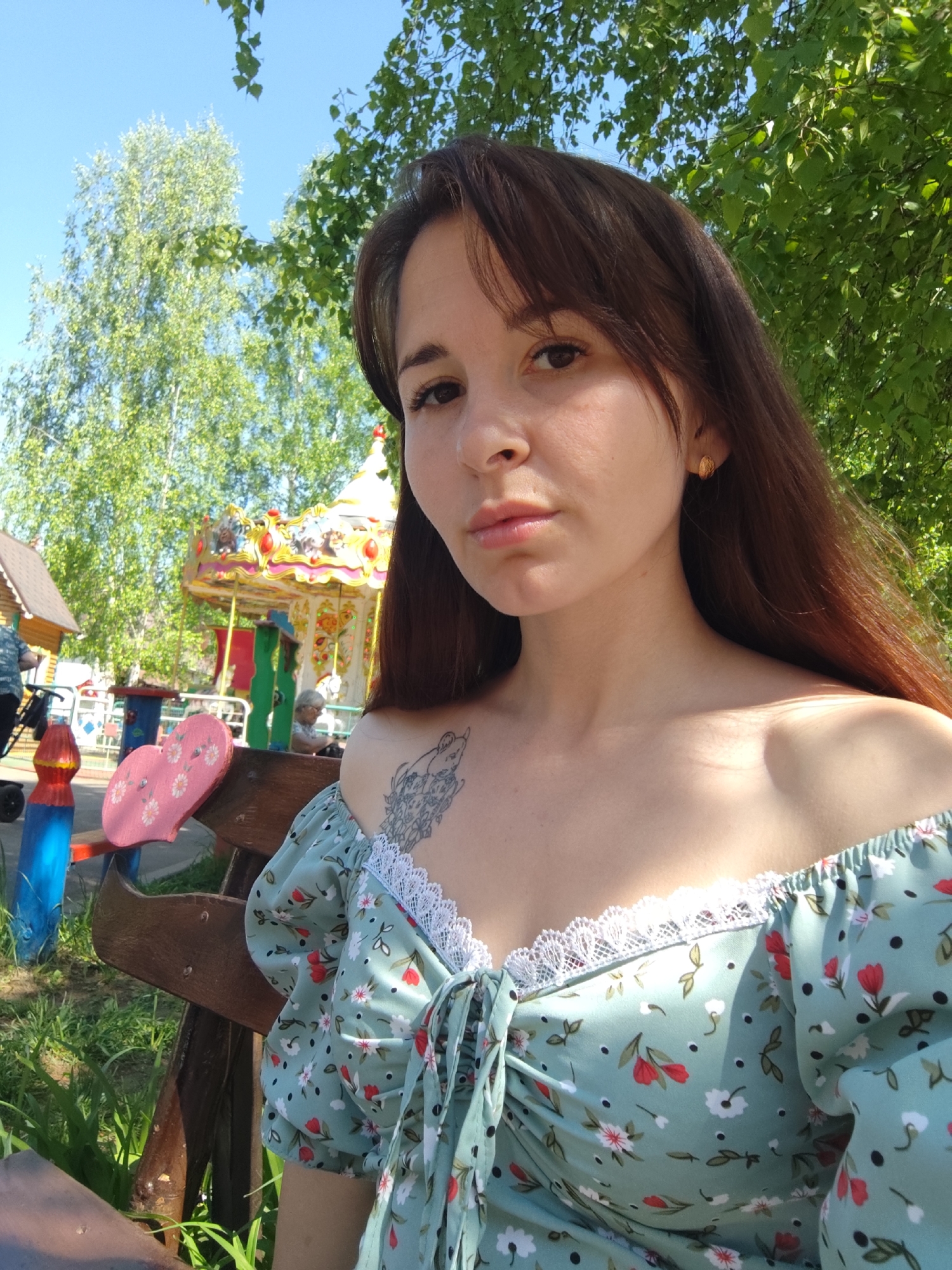 A woman stands outdoors in Chaikovsky, Russia, in front of a carousel and a small house. She is fair-skinned with brown hair and is wearing a floral, off-the-shoulder dress with white lace trim. A tattoo is visible on her upper chest. She is sitting on a bench, and a portion of a table can be seen. In the background, there is a colorful carousel, a small wooden house, and trees.