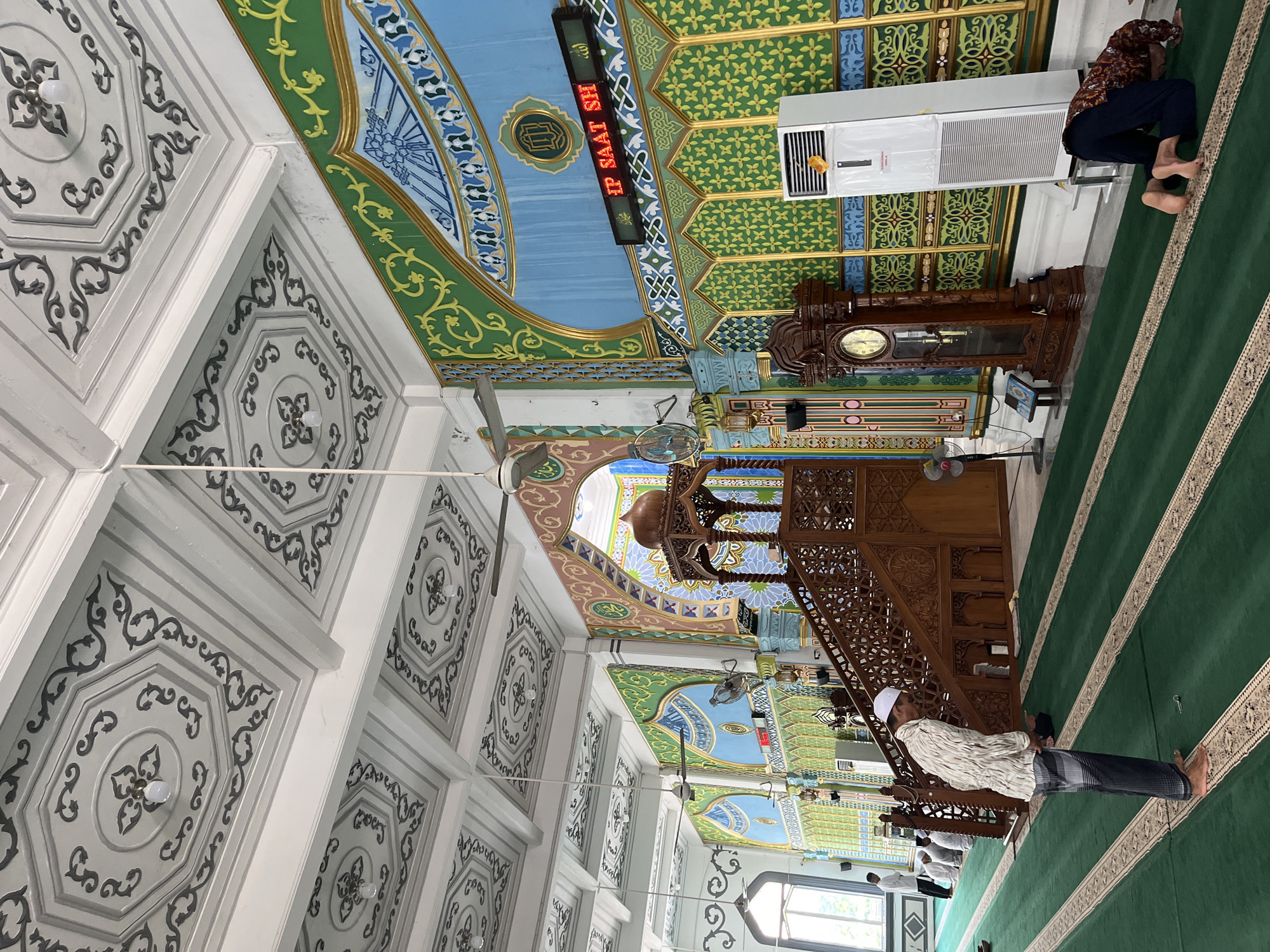The interior of a mosque in Keudekruenggeukueh, Indonesia, displays elaborate ceiling decorations in white, green, blue, and gold colors. A multi-tiered wooden minbar stands prominently. An LED display indicates the time. A standing grandfather clock is also present. Individuals are observed praying on green carpets. An air conditioning unit is visible on the wall.