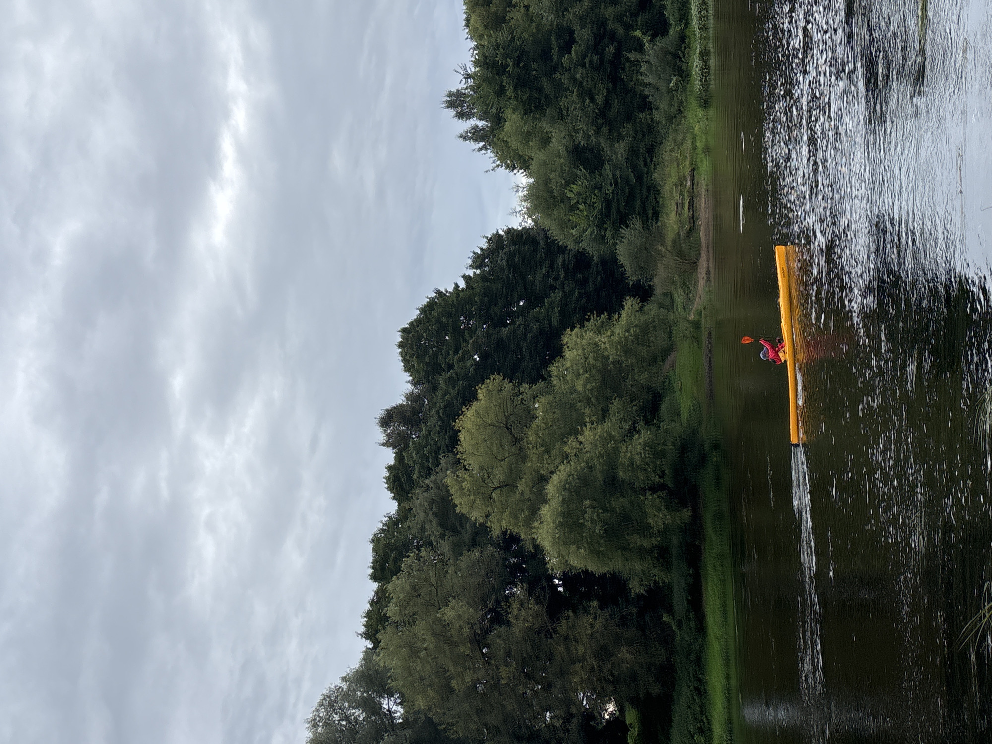 A person in a yellow kayak paddles on a wide, dark river. The kayaker is wearing a red jacket and is visible from the chest up. The kayak leaves a faint wake. The riverbank is lined with lush green trees. The sky is overcast with gray clouds. The scene is likely located in the city of Zvenigorod, Russia, given the natural landscape and implied setting.