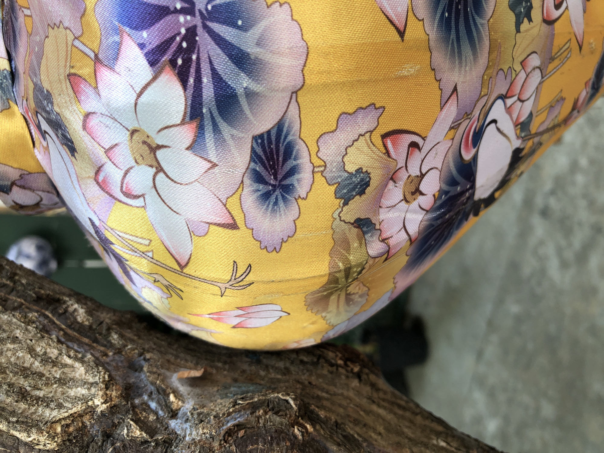 The image shows a close-up of a yellow fabric with a floral pattern, likely a textile. The fabric is draped over a rough, textured surface, possibly a tree branch or driftwood, which occupies the foreground. The floral design features large, stylized flowers in shades of pink, white, blue, and purple, with intricate details and outlines. Some of these flowers appear to be lotuses or similar blooms, rendered in a traditional East Asian art style. The fabric's surface has a slight sheen, suggesting it might be silk or a similar material.
In the background, a blurred green surface is visible, possibly foliage or a painted wall, and beyond that, a muted gray, textured background, which could be a concrete floor or wall. There are hints of other objects in the background, including a small, out-of-focus sphere that could be a decorative item or a person's head. The lighting suggests an indoor or shaded outdoor setting, with no strong shadows indicating direct sunlight.
The image does not depict any people engaged in an activity, nor does it capture a specific event. The focus is entirely on the texture and pattern of the fabric and its juxtaposition with the natural element in the foreground. No text is visible in the image. The setting cues, such as the rustic foreground and the possibility of an East Asian artistic style in the fabric pattern, combined with the provided location context of Đà Nẵng, Vietnam, might suggest this is an object within a Vietnamese home, shop, or garden.