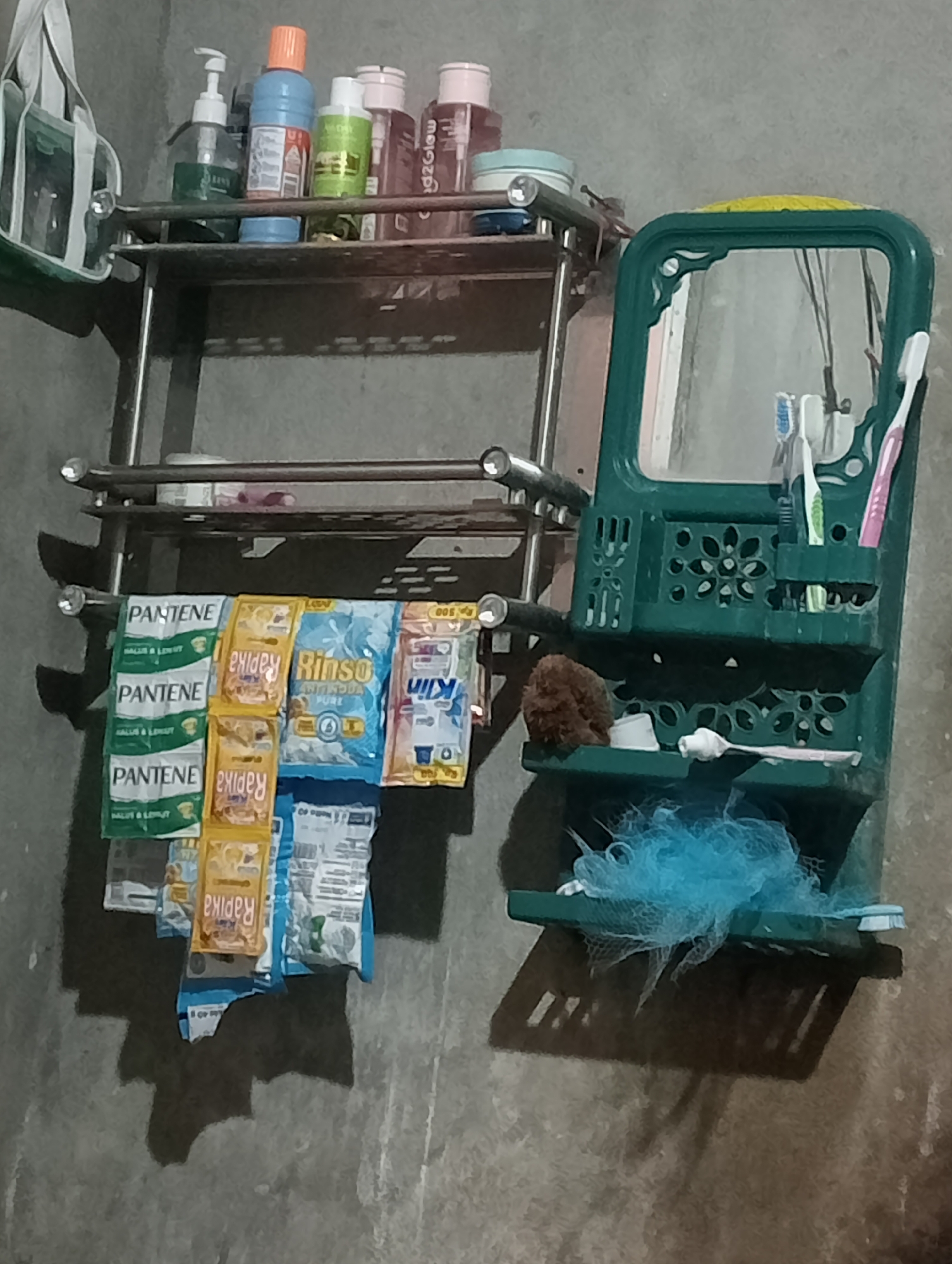 An indoor scene features a shelf with various toiletries and personal care items, along with a toothbrush holder with a mirror. The wall behind is gray and textured. On the top shelf, bottles of shampoo and conditioner are visible. Below, hanging from a rack, are sachets of Pantene shampoo, Rapika hair vitamins, Rinso detergent, and Kin toothpaste. To the right, a green plastic toothbrush holder with compartments holds toothbrushes, a small brush, and a bar of soap. A blue loofah is placed on the bottom shelf of the holder. The overall impression is of a functional bathroom storage area.