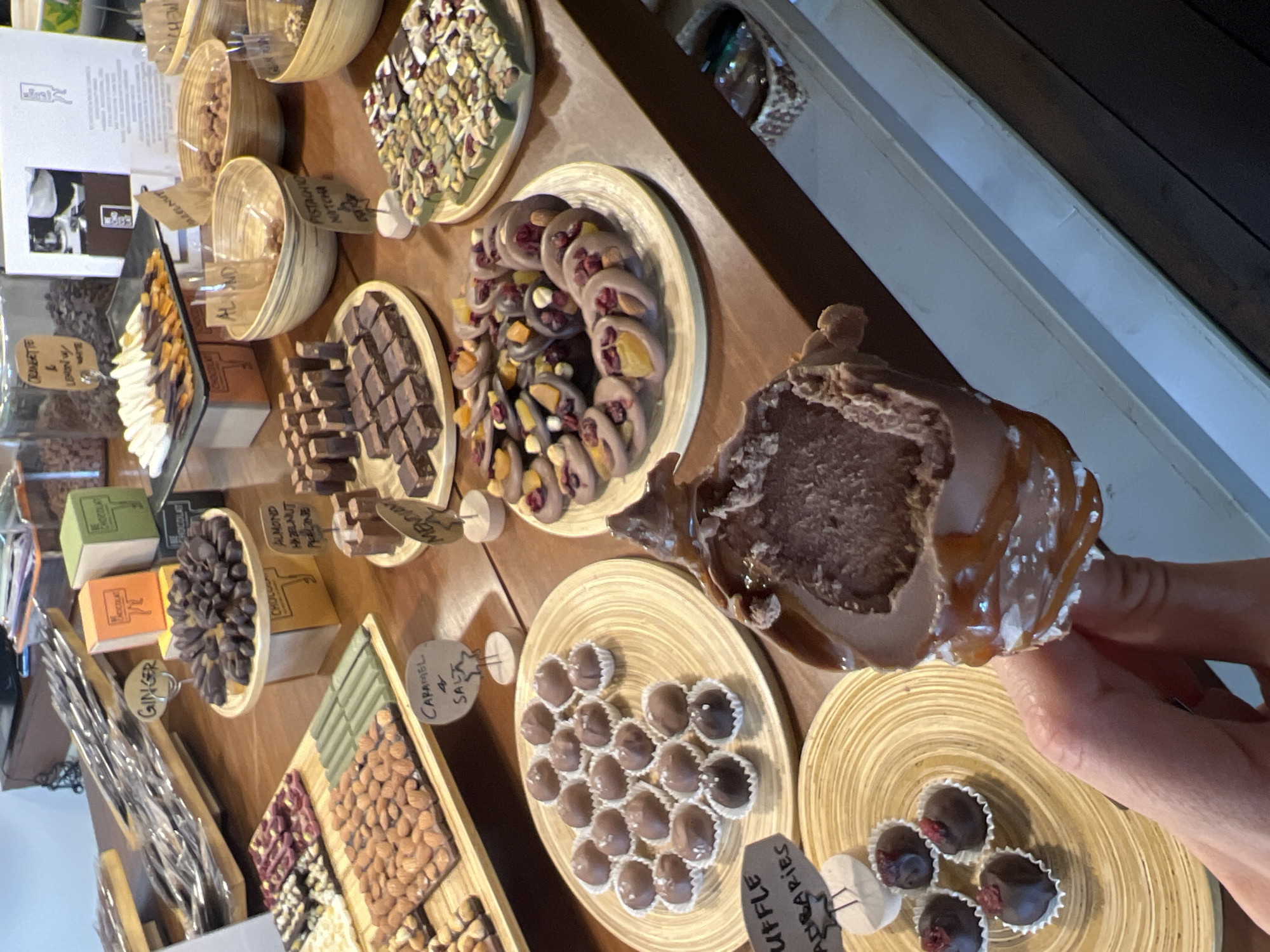 The image shows a display of chocolates in an unknown city, at an unknown location. Various types of chocolates are arranged on wooden plates and shelves, including truffles, bars, and other confections. Some of the chocolates are labeled with descriptions such as "Caramel & Salt," "Almond Hazelnut Praline," "Ginger," "Cranberry & Lemon," and "Pistachio Matcha." A hand is holding a large chocolate item, with a visible cross-section. The environment is a retail setting, likely a chocolate shop or confectionery.