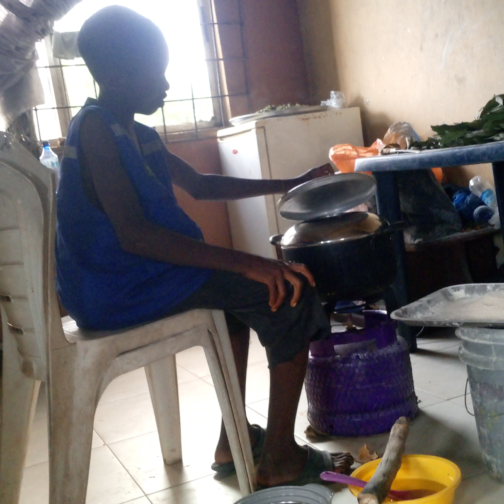 A side-profile shot captures a person, likely a child or young individual, with dark skin, seated on a white plastic chair in what appears to be a kitchen or utility room. The person wears a blue sleeveless top and dark shorts, and is looking towards the right side of the frame, with their right hand positioned near a large, dark cooking pot. Another smaller pot or lid is stacked on top of the first cooking pot. This setup suggests the person is either engaged in cooking or observing the process.
In the background, bright daylight streams through a window fitted with metal grilles, indicating it is daytime. A white refrigerator stands against a light brown wall, with leafy greens on a tray placed on its top. To the right of the frame, a blue plastic table holds various items, including plastic bottles and possibly more greens.
On the tiled floor, near the person's feet, are grey buckets, a distinctive purple gas cylinder (commonly used for cooking stoves), and a yellow bowl containing a brown wooden stick and a purple utensil, likely used for stirring or serving. The overall setting is a functional indoor space, possibly a home, centered around food preparation. No discernible text is visible in the image.