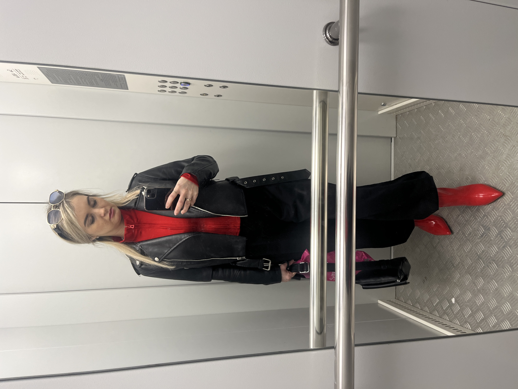 A young woman is taking a selfie in an elevator with white walls and a steel-plated floor. She is wearing a red top, black pants, a black leather jacket, and red pointed-toe boots. Sunglasses are perched on her head. She is holding a black purse and a pink shopping bag. The elevator features a control panel and metal handrails. The location is Chaikovsky, Russia.