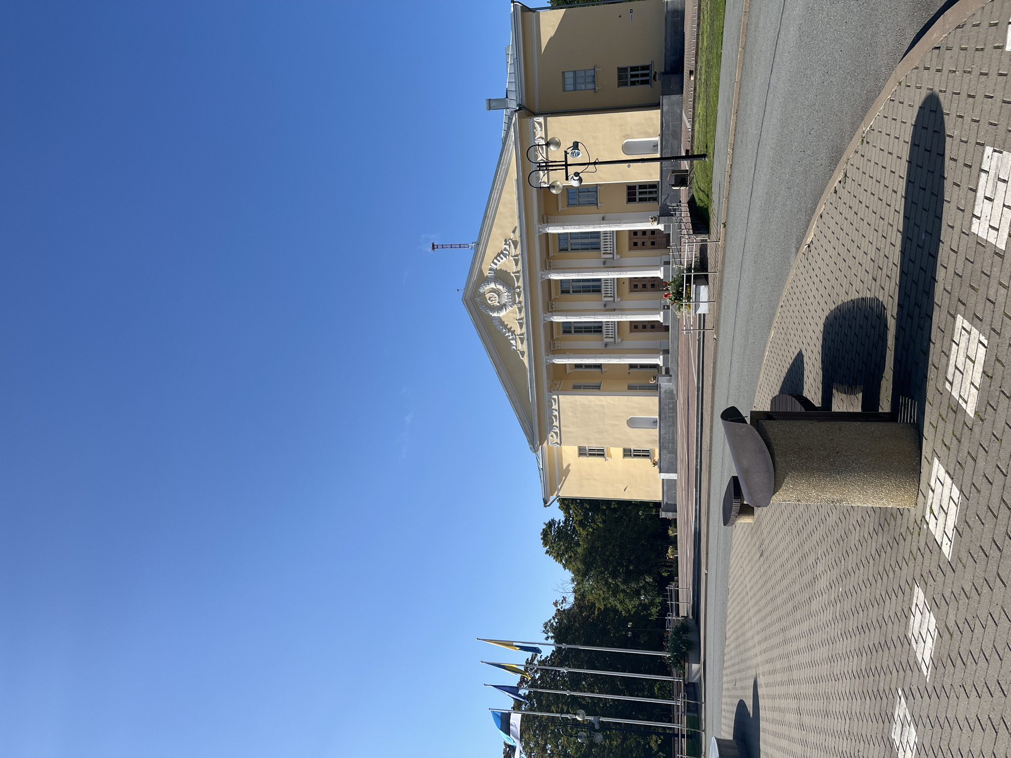 The image captures a wide-angle, low-angle view of a light yellow, neoclassical building with a prominent white portico supported by columns. The building, identified as the Kohtla-Järve Cultural Centre, is situated in the city of Kohtla-Järve, Estonia. A bright blue sky with no visible clouds forms the backdrop.
In the foreground, a paved area with a brick-like pattern is visible, featuring some white square inlays. A curved asphalt road is to the right of the paved area. A concrete trash receptacle with a dark metal lid and a wooden bench are positioned on the paved area.
To the left of the building, four flagpoles stand tall, flying the Estonian flag, a blue flag, and two other flags, one yellow and blue, and another mostly yellow. Behind the flags, a dense line of green trees creates a backdrop for the building.
The building itself has a symmetrical facade with multiple windows and arched niches. A decorative relief is centered above the main entrance. A street lamp with multiple lights stands in front of the building on the right side. The scene is well-lit, suggesting it was taken during daylight, likely in the morning or afternoon, given the clear sky and shadows. There are no people visible in the image. The overall impression is one of a well-maintained public space on a clear day.