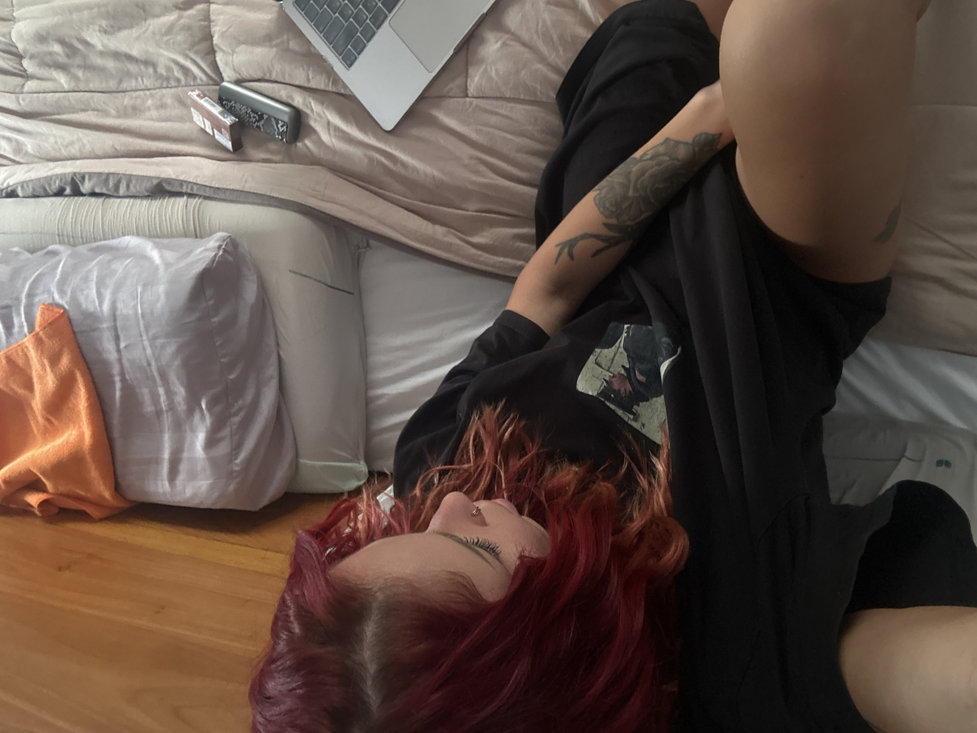 A person with dyed red hair is lying on a bed indoors. The person is wearing black clothing and has a tattoo on their left arm. A laptop and other personal items are on the bed next to them. The bed is covered with a beige duvet and pillows. A wooden floor is visible. There is no indication the image was taken in North Kuta, Indonesia.