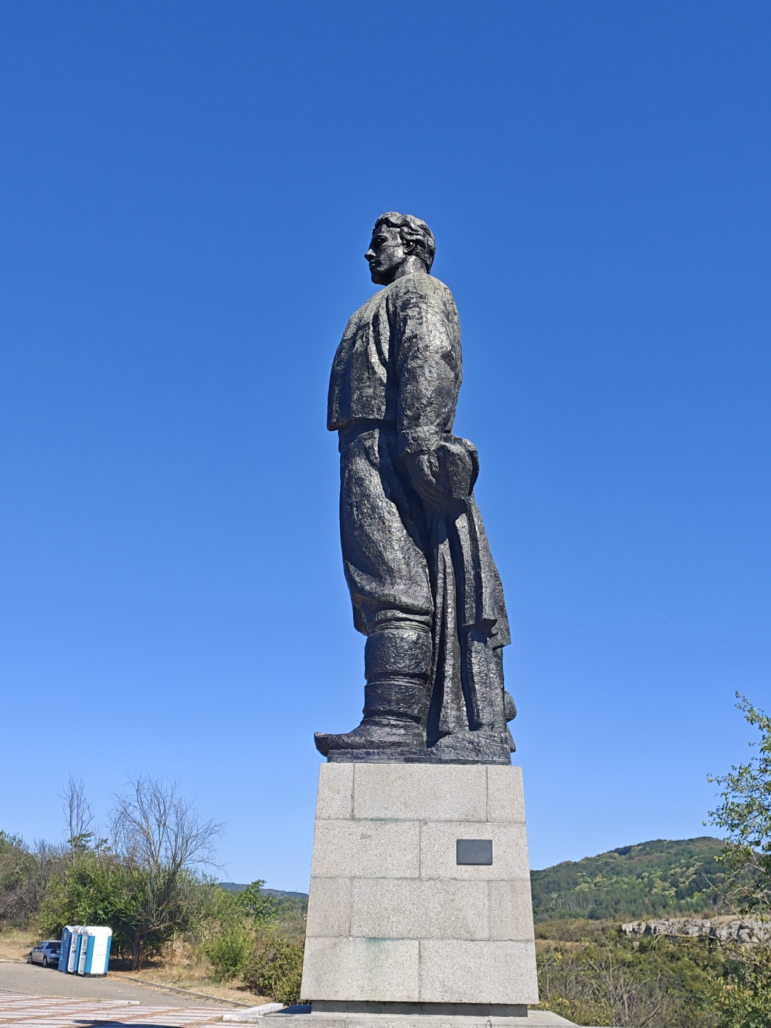 The image shows a tall, dark bronze statue of a man in historical attire, standing on a rectangular stone pedestal. The statue is positioned in profile, facing left. The man is wearing what appears to be a jacket, trousers tucked into boots, and a cloak or overcoat draped over his arm. His hair is styled in waves. The pedestal is made of large, light-colored stone blocks and has a dark plaque on its front.
The statue is situated outdoors with a clear, bright blue sky in the background. In the mid-ground and background, there are hills covered with green trees and scrub. To the left of the pedestal, at the base of the monument, a paved area is visible, with a silver car parked next to three blue and white portable toilets. The overall environment suggests an open, possibly historical or memorial site.
This statue is located in the city of Lovech, Bulgaria, and it is identified as the monument to Vasil Levski. Vasil Levski was a prominent Bulgarian revolutionary who fought for the liberation of Bulgaria from Ottoman rule. The monument is situated on a hill overlooking the city.
