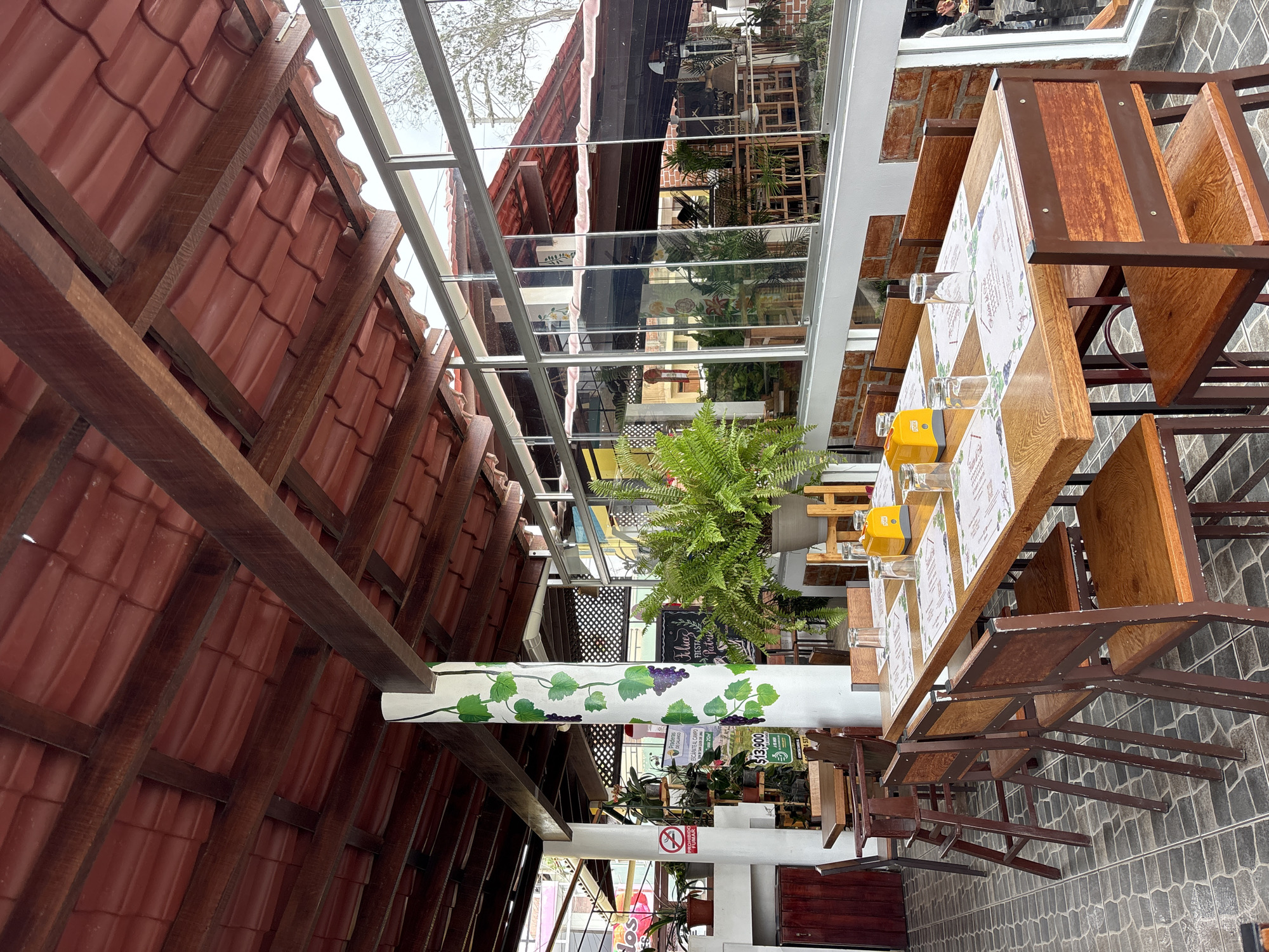 The image shows an outdoor dining area of a restaurant in San Vicente de Azpitia, Peru. The scene is set under a reddish-brown tiled roof supported by wooden beams. A large white pillar, decorated with painted green ivy and purple grapes, stands prominently in the foreground. Several wooden tables are set with placemats, glasses, and yellow dispensers. Wooden chairs are arranged around the tables. Large glass windows line the side of the restaurant, offering a view of the interior and the street beyond. Lush green ferns in a pot sit on a wooden stand. A "No Smoking" sign is visible on a white wall to the left. The flooring is made of grey, patterned tiles. The overall impression is of a casual, open-air dining establishment.
