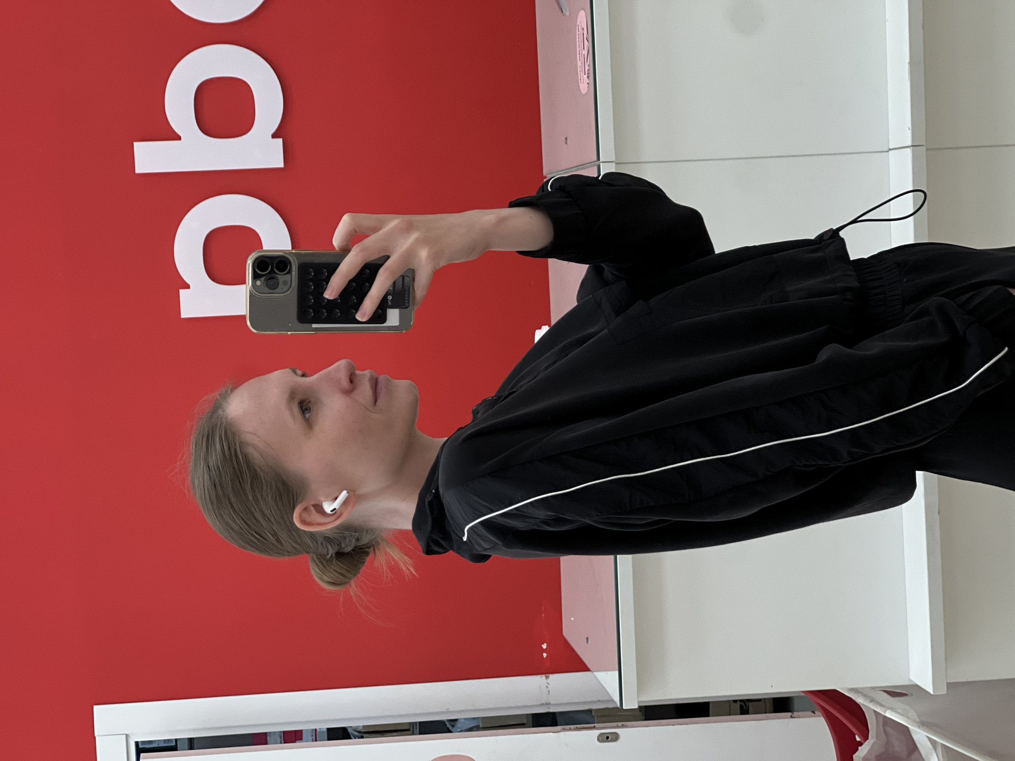 A young woman with light brown hair pulled back into a bun stands in front of a bright red wall with large white lettering. She holds a smartphone with a dark case in her right hand, presumably taking a photo or video. She wears an Apple AirPod in her left ear and is dressed in a black tracksuit with a white stripe along the sleeve. The background also includes a white countertop or shelf and hints of a white wall or structure on the right. It is unknown whether this image was taken in Khimki, Russia.
