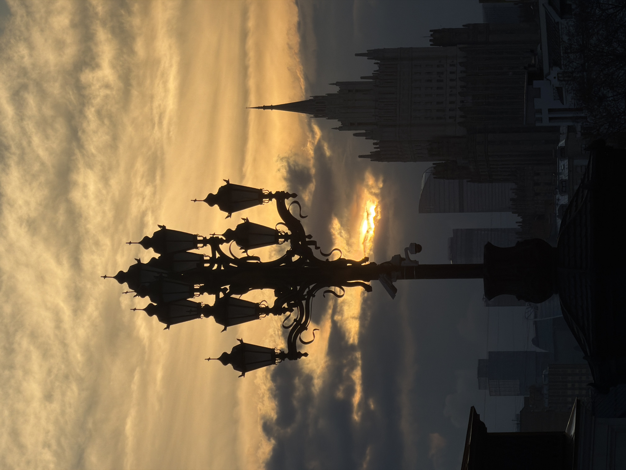 The image captures a view of Moscow, Russia at sunset. The silhouette of an elaborate lamppost dominates the foreground, contrasting with the vibrant orange and yellow sky. Behind the lamppost, the cityscape is partially obscured by the fading light and atmospheric conditions. Prominently featured is the Moscow State University main building, identifiable by its spire. Various other modern buildings are also discernible in the background.