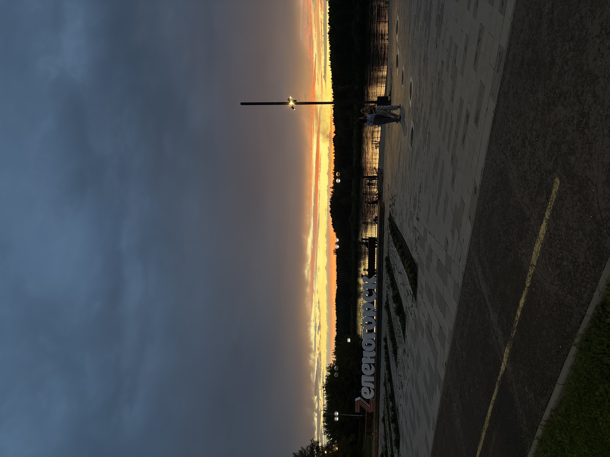 The image depicts a waterfront promenade in Zelenogorsk, Russia, during a sunset. In the foreground, a paved area with geometric patterns leads towards the water. To the left, large illuminated white letters spell out "Zelenogorsk." In the mid-ground, a river flows, reflecting the warm hues of the sunset. A dark tree line borders the opposite bank of the river. Two individuals are visible walking on the promenade in the mid-ground, silhouetted against the sunset. A tall lamp post with lights stands near them. The sky is dramatic, with dark clouds juxtaposed against vibrant streaks of orange and yellow from the setting sun. The overall atmosphere is serene and picturesque, capturing a moment of natural beauty in an urban setting.