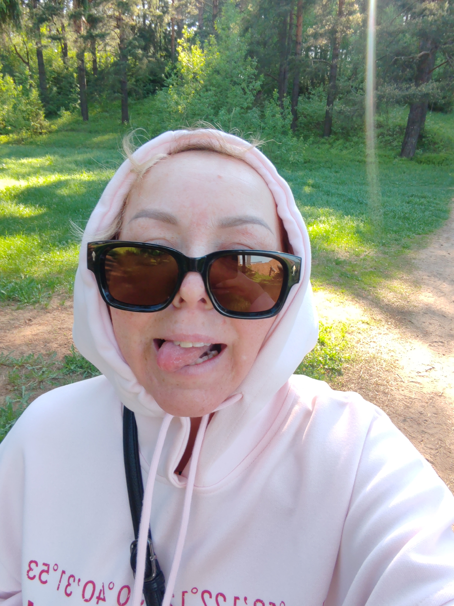A woman with fair skin is taking a selfie in a park. She is wearing a light pink hoodie and oversized black sunglasses. Her tongue is sticking out, and she is smiling. A black purse strap is visible on her left shoulder. The park has trees and green grass. The photograph was taken during daytime under bright sunlight. The text printed on the hoodie is not clearly readable due to being upside down. Based on the text on the hoodie and visual information, it can be inferred that the location of the photograph is Chaikovsky, Russia.