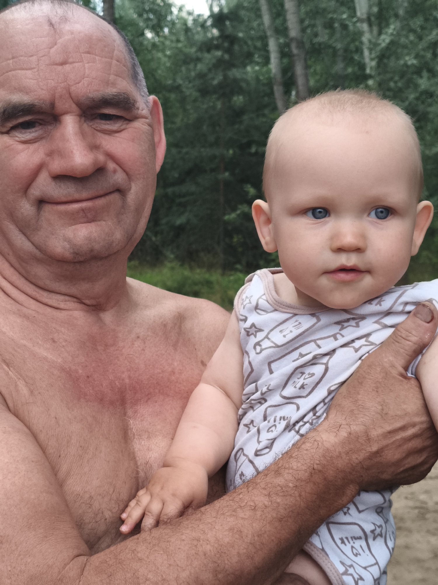 A man and a baby are in the foreground, with a blurry background of trees. The man, who is shirtless, is holding the baby. The baby is wearing a light blue onesie with a pattern of milk bottles and stars. Both the man and the baby have blue eyes. The man is smiling slightly, and the baby is looking towards the camera. The lighting suggests it is daytime, possibly outdoors in a natural setting.