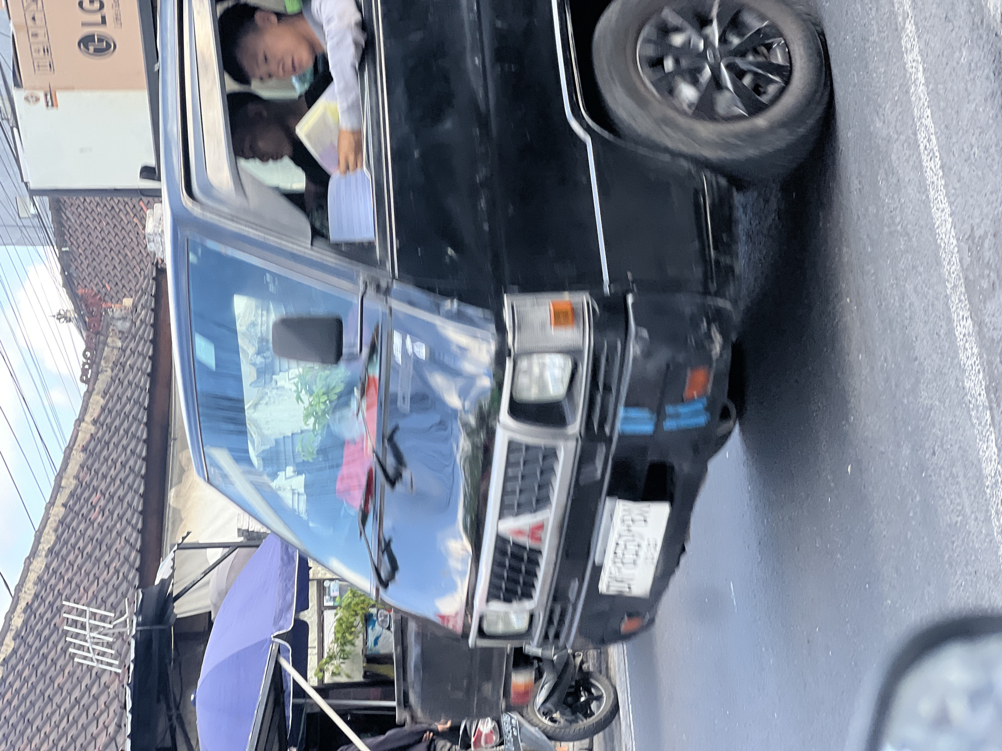 A black Mitsubishi L300 van is captured in motion on a city street in Denpasar, Indonesia. Two young men are visible inside the van. The driver, positioned on the right side of the vehicle, is partially obscured by the frame. The passenger on the left is leaning forward, holding a stack of papers. The van's front grille features the Mitsubishi logo. In the background, a blue umbrella and a tiled roof building are visible. The street surface is asphalt with white lane markings. A portion of a motorcycle is also visible on the left side of the frame, suggesting it is in motion alongside the van. The overall scene depicts a typical urban street environment.
