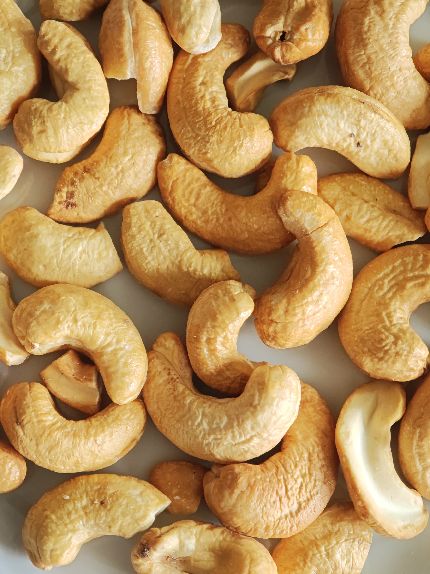 The image is a close-up, overhead shot of a pile of roasted cashew nuts. The nuts are a golden brown color and are scattered across a light grey surface. Some of the cashews are whole, while others are broken into pieces. The texture of the cashews appears slightly rough and bumpy. There are no people or animals present in the image. The lighting is even, casting minimal shadows, suggesting an indoor setting. The specific location in the city of Kursk, Russia, cannot be determined from the visual information provided.
