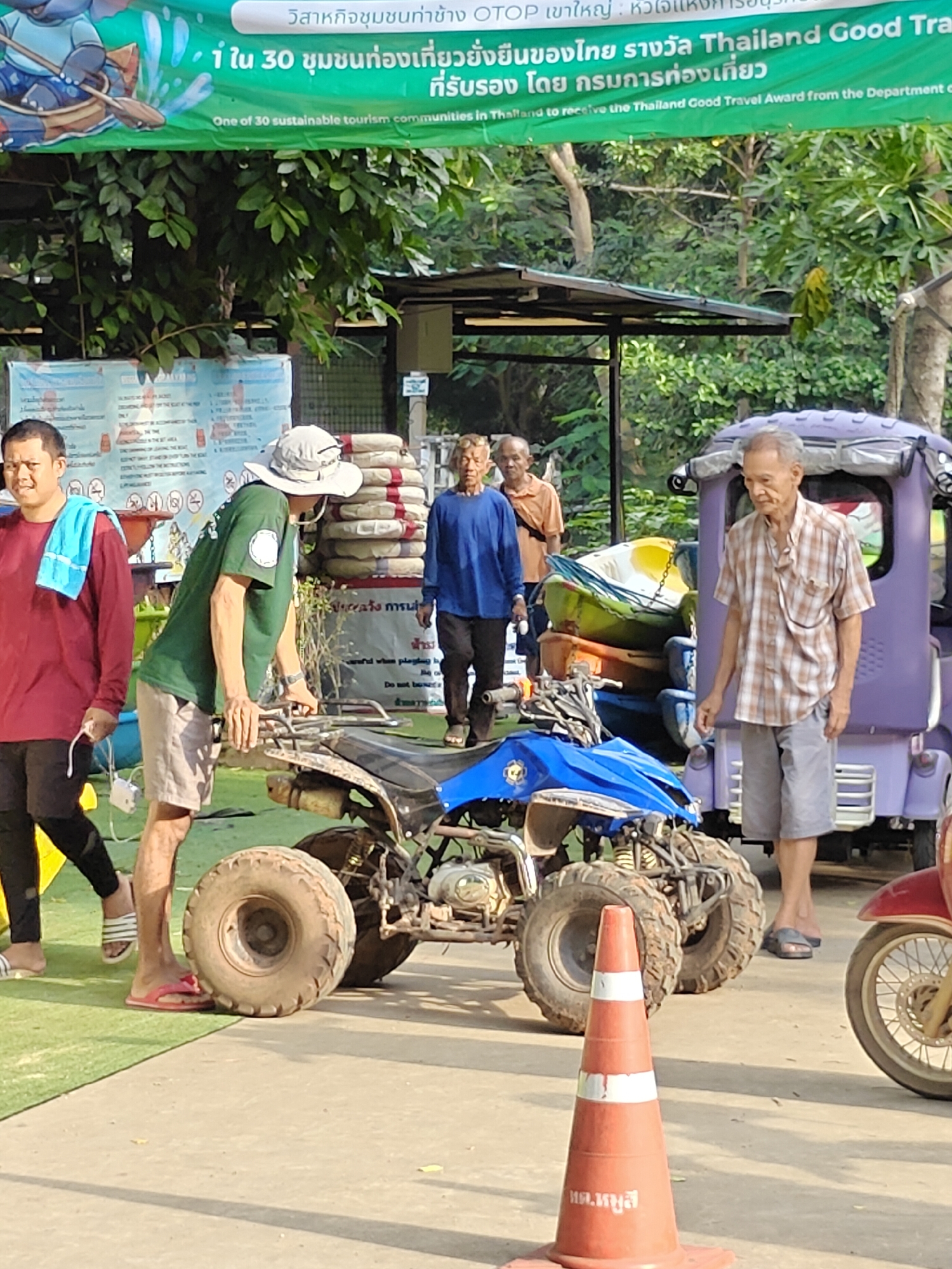 The image depicts an outdoor scene, likely in Tambon Mu Si, Thailand. In the foreground, a blue all-terrain vehicle (ATV) is parked on a concrete path adjacent to a grassy area. A traffic cone with Thai writing is positioned in front of the ATV. To the left of the ATV, a person wearing a maroon long-sleeved shirt and black pants stands with a blue towel draped over their shoulder. Next to them, another person in a green long-sleeved shirt and beige shorts is leaning on the ATV's handlebars, wearing a wide-brimmed hat. Further back, several older men are visible. One man in a blue long-sleeved shirt stands near a stack of life preservers. Another man in an orange shirt is also present. To the right of the ATV, a purple vehicle resembling a tuk-tuk is parked, and an elderly man in a plaid shirt and grey shorts walks away from it. A red motorcycle is partially visible on the far right of the image. A banner with Thai and English text is hung in the background, suggesting a promotional or informational display. The environment appears to be a recreational or tourist area, with trees and foliage visible in the background.