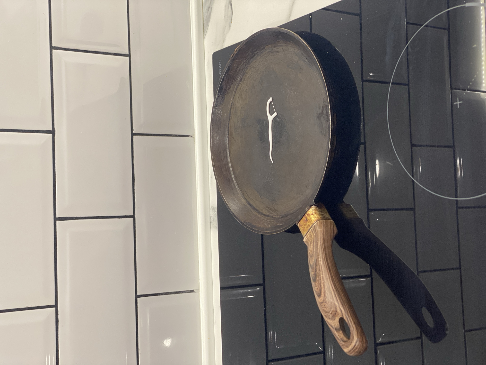 A white plastic toothpick is resting inside a dark, round frying pan on a black induction stovetop. The frying pan has a wooden handle with a brass-colored metal attachment to the pan. The stovetop is set against a backsplash of white subway tiles with black grout. The scene is lit from above, casting a shadow of the pan and handle onto the stovetop. The location is identified as Avtozapchasti, Russia, though this is likely a misinterpretation as the visual elements do not directly suggest this location.