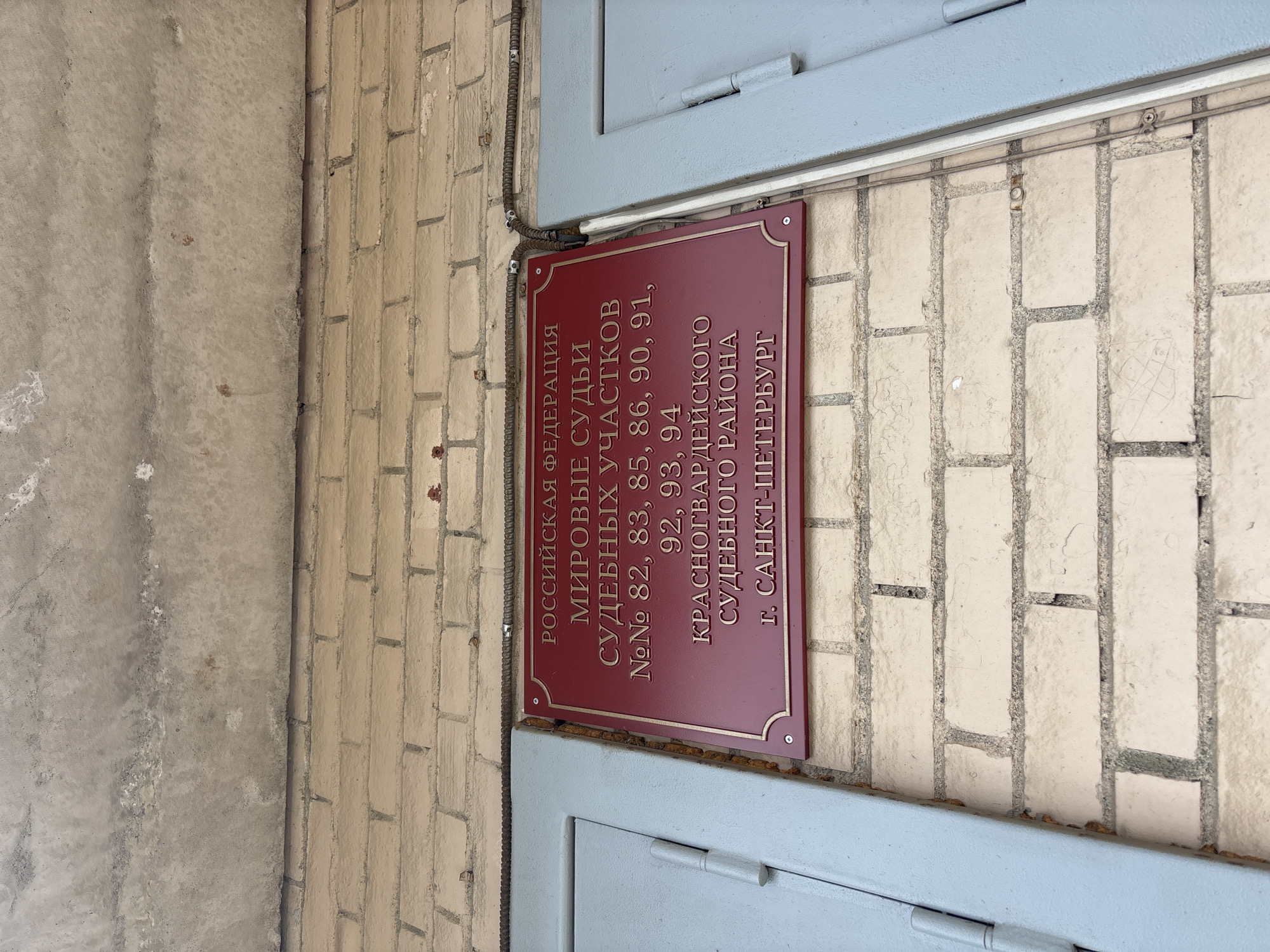 The image shows a sign on a brick wall. The sign is red with gold lettering in Russian, indicating that the location is a court of justice for various districts. Specifically, it denotes the court of justices of the peace for court districts 82, 83, 85, 86, 90, 91, 92, 93, and 94, located in the Krasnogvardeysky District of the city of Saint Petersburg, Russia. The sign is bordered in gold and secured to the wall with screws. Part of a metal doorframe is visible around the sign.