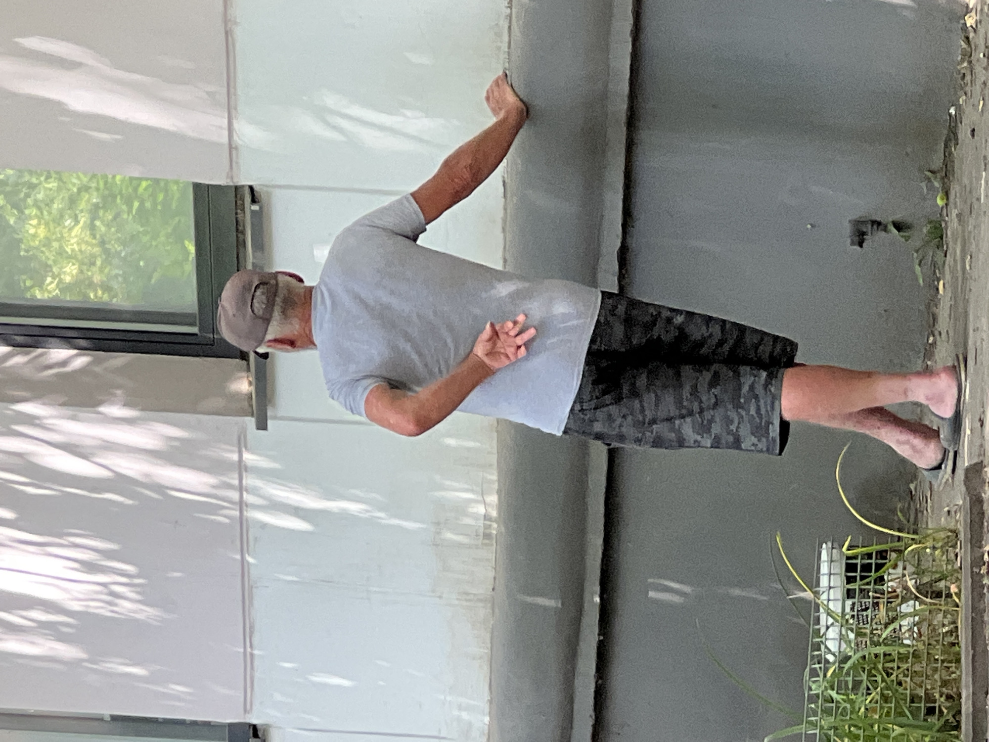 A man wearing a grey t-shirt, camouflage shorts, a baseball cap, and flip-flops leans against a building wall. His right arm is extended and resting on the wall, while his left arm is bent with his hand behind his back. He is facing away from the camera, looking towards the right side of the frame. The background consists of a building with a window showing green foliage outside and shadows cast on the wall from trees. In the foreground, there is a metal fence with some greenery growing inside and around it, and the ground is paved with what appears to be concrete and dirt. This scene is likely outdoors in a city setting.