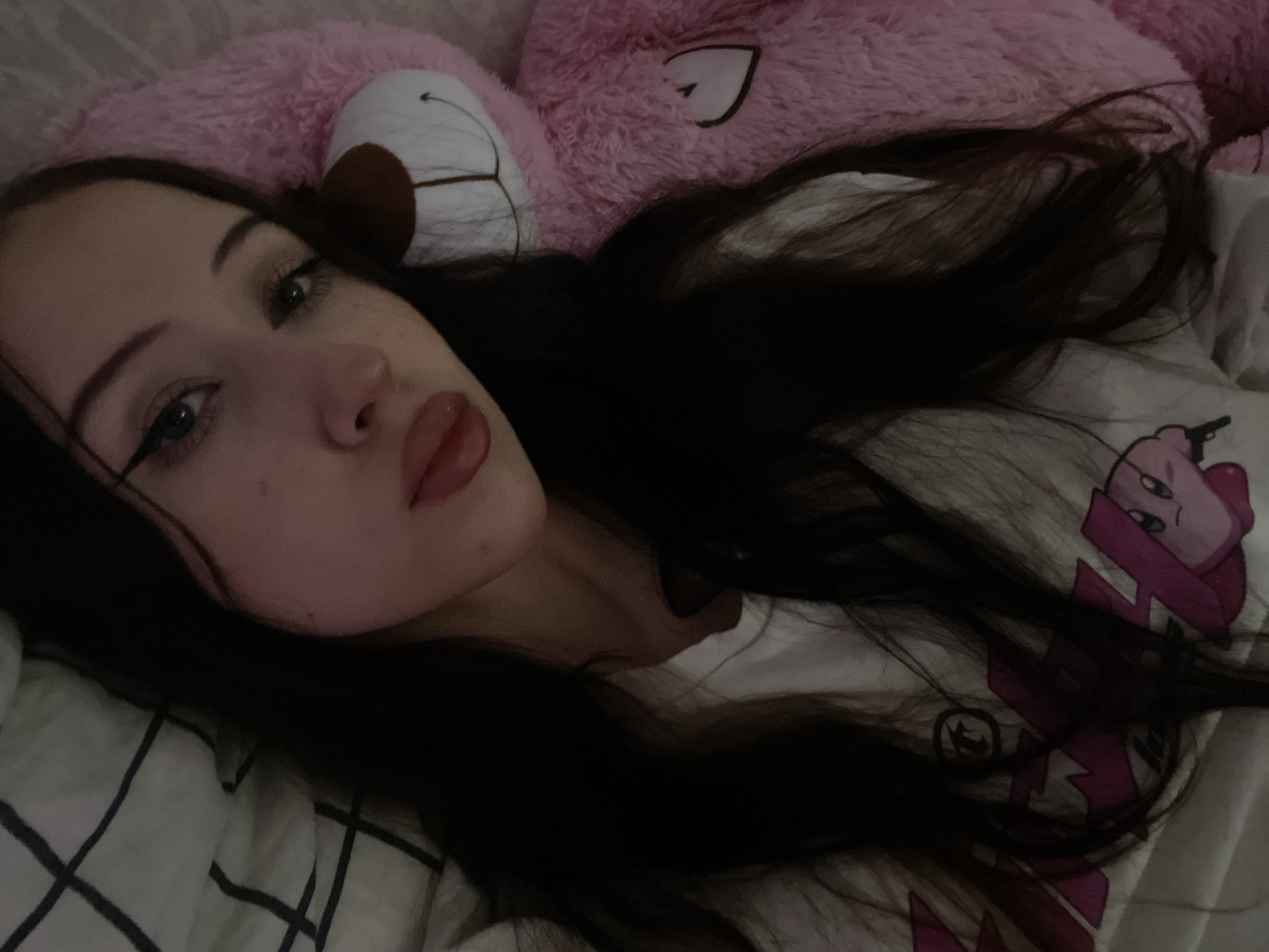 In an indoor setting, likely a bedroom, a young woman with long, dark hair lies down next to a large, pink teddy bear. She is wearing a white t-shirt with a graphic print featuring characters from the video game "Kirby." The woman's face is partially visible, showing her eyes with winged eyeliner and her lips with lipstick. The teddy bear is positioned behind her, its head and torso visible. The background includes a white and dark-lined patterned sheet and a portion of the pink teddy bear. The image appears to be a selfie taken from a close-up perspective, focusing on the woman and the teddy bear. There is no information in the image to suggest a location in Votkinsk, Russia, or any other specific city.