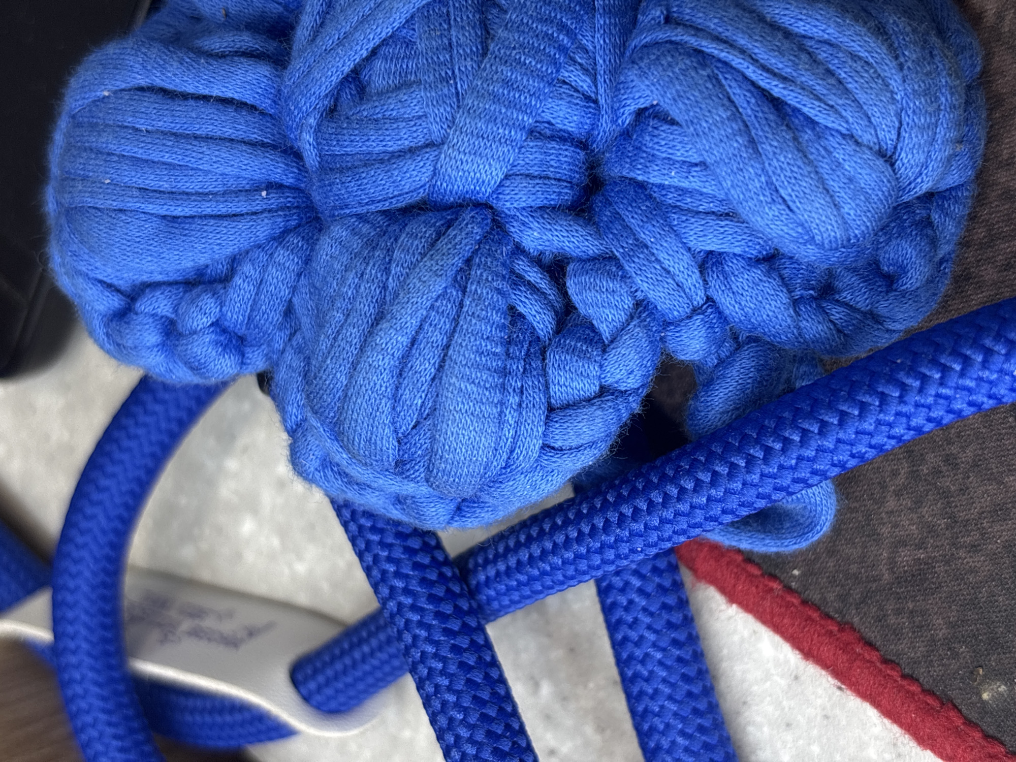 A close-up shows a bright blue knitted or woven object, possibly a hat or decorative element. In the foreground, two rounded blue cords overlap, intersecting with the edge of a white material with a red stripe, and a section of grey material. The image appears to be taken indoors under relatively bright lighting conditions. There is no evidence suggesting the location is in Perm, Russia.