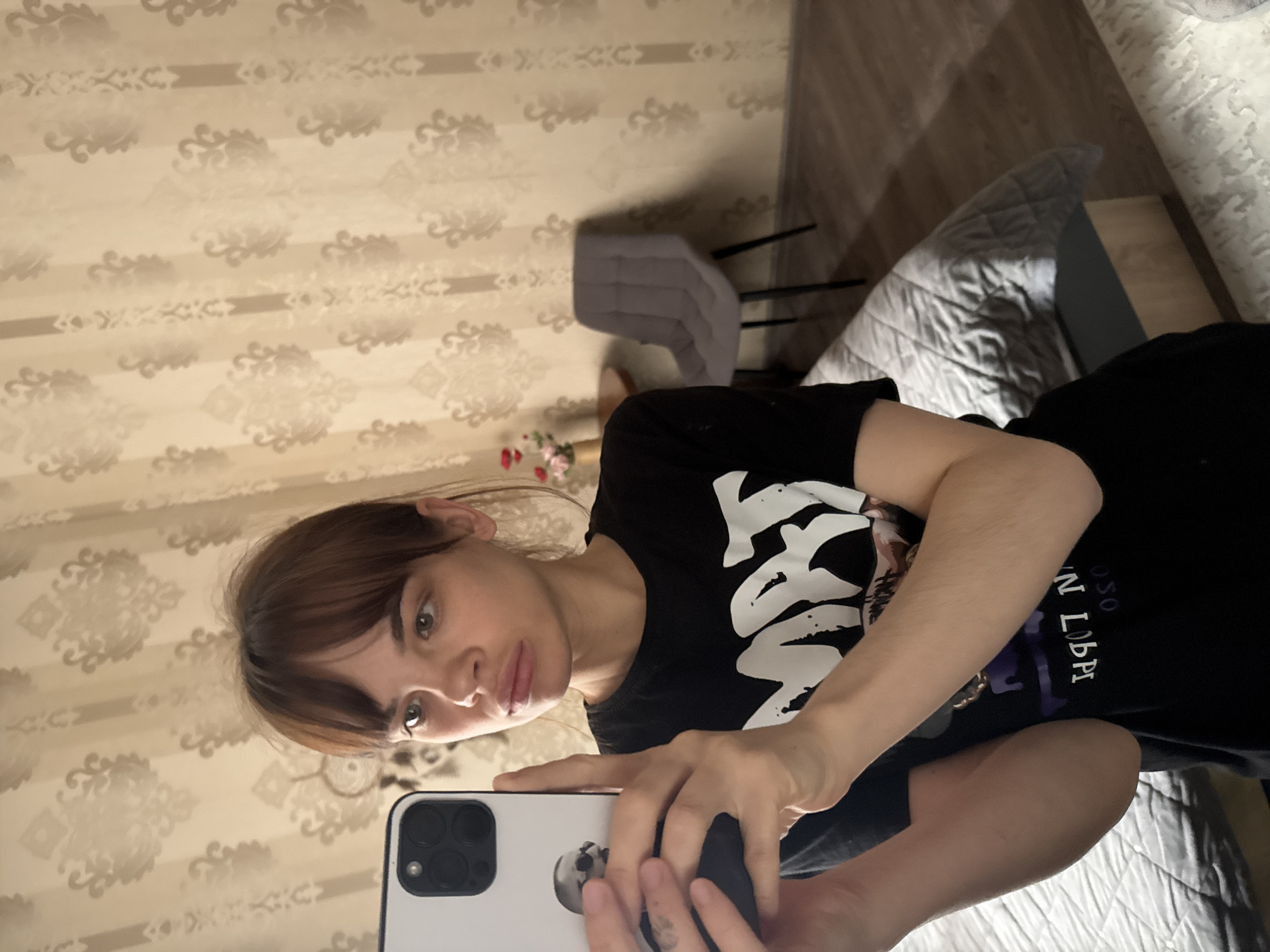 The image depicts a young woman taking a mirror selfie in what appears to be a residential indoor setting, likely a bedroom or living space in Rostov-on-Don, Russia. She is positioned to capture her upper body, holding a white smartphone, likely an iPhone, with her right hand. The phone's multi-lens camera system is visible, and its case features a skull design.
The woman has light brown hair with bangs, fair skin, and a neutral, slightly pensive expression as she looks into the phone. She is wearing a black t-shirt with a prominent white graphic design on the front. Visible text on the shirt, though stylized, includes what appears to be "OSON LBPI".
The background features light-colored wallpaper adorned with a repeating, ornate damask-like pattern. The floor is covered with light wood laminate or paneling. A gray upholstered chair with black legs is visible behind her. To the right, a bed is partially visible with a light gray quilted comforter. A small vase containing pink flowers is situated on a surface in the lower-left portion of the background. The lighting suggests it is daytime indoors, though no specific time or weather can be determined. The scene conveys a personal and relaxed atmosphere.