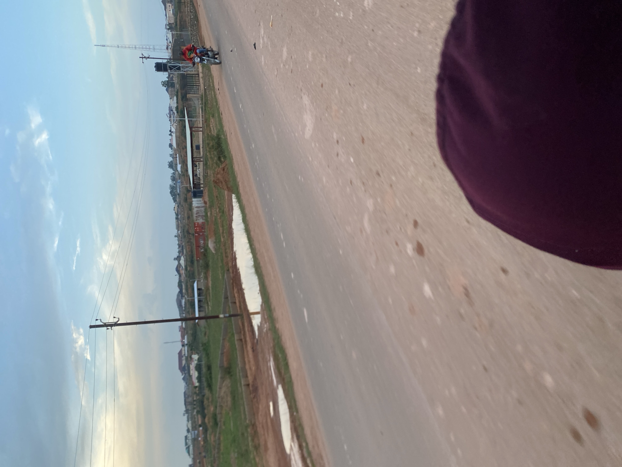 The image shows a view of a dusty, unpaved road in Kwang, Nigeria, with a person on a motorcycle in the distance. The sky is cloudy, and there are houses and trees in the background. The foreground is out of focus, showing what appears to be the shoulder or back of a person's head. The road is lined with dry grass and some puddles of water. A utility pole with wires runs along the side of the road. The overall impression is of a rural or semi-rural setting on a slightly overcast day.