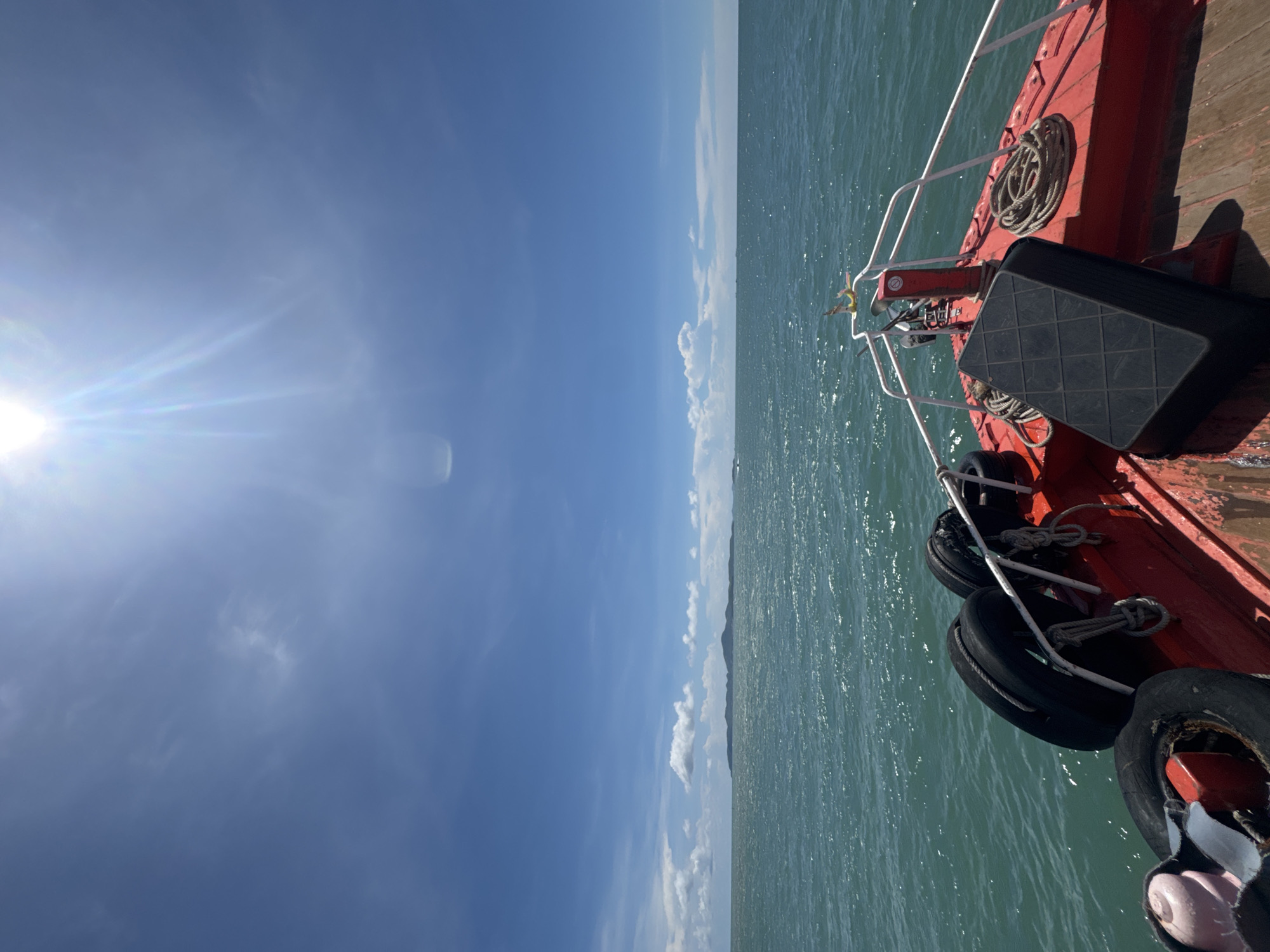The image shows a view from a boat in Pattaya City, Thailand. The sea is on the right side of the image, while a blue sky with the sun is on the left. A portion of a red boat with white railings is visible on the right edge. Several tires and a black rectangular object are attached to the side of the boat. The water has a greenish tint and slight waves. In the distance, a faint outline of land can be seen along the horizon. The sky is mostly clear with a few scattered clouds.