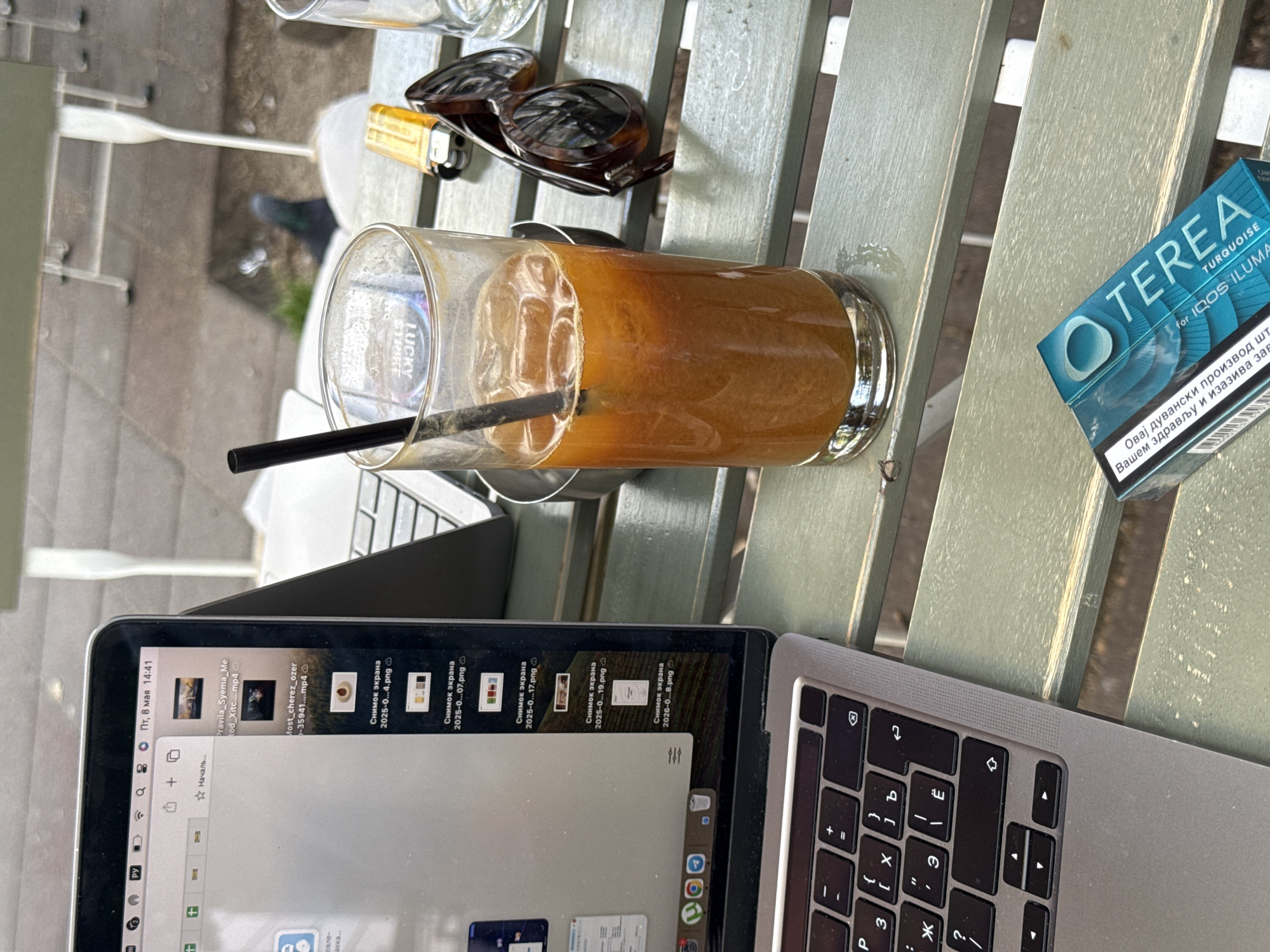A table on an outdoor patio features a laptop with a file explorer window open and a Cyrillic keyboard, a glass of iced coffee with a black straw, a pack of Terea Turquoise for IQOS Iluma cigarettes, sunglasses, and a lighter. The laptop's display shows files named "Снимок экрана" followed by dates in 2025 and 2026, as well as filenames containing "Xitc", "ozer", and "35941". The cigarette pack has text in Serbian, indicating the city is likely Belgrade, Serbia. The patio furniture appears to be green and slatted.