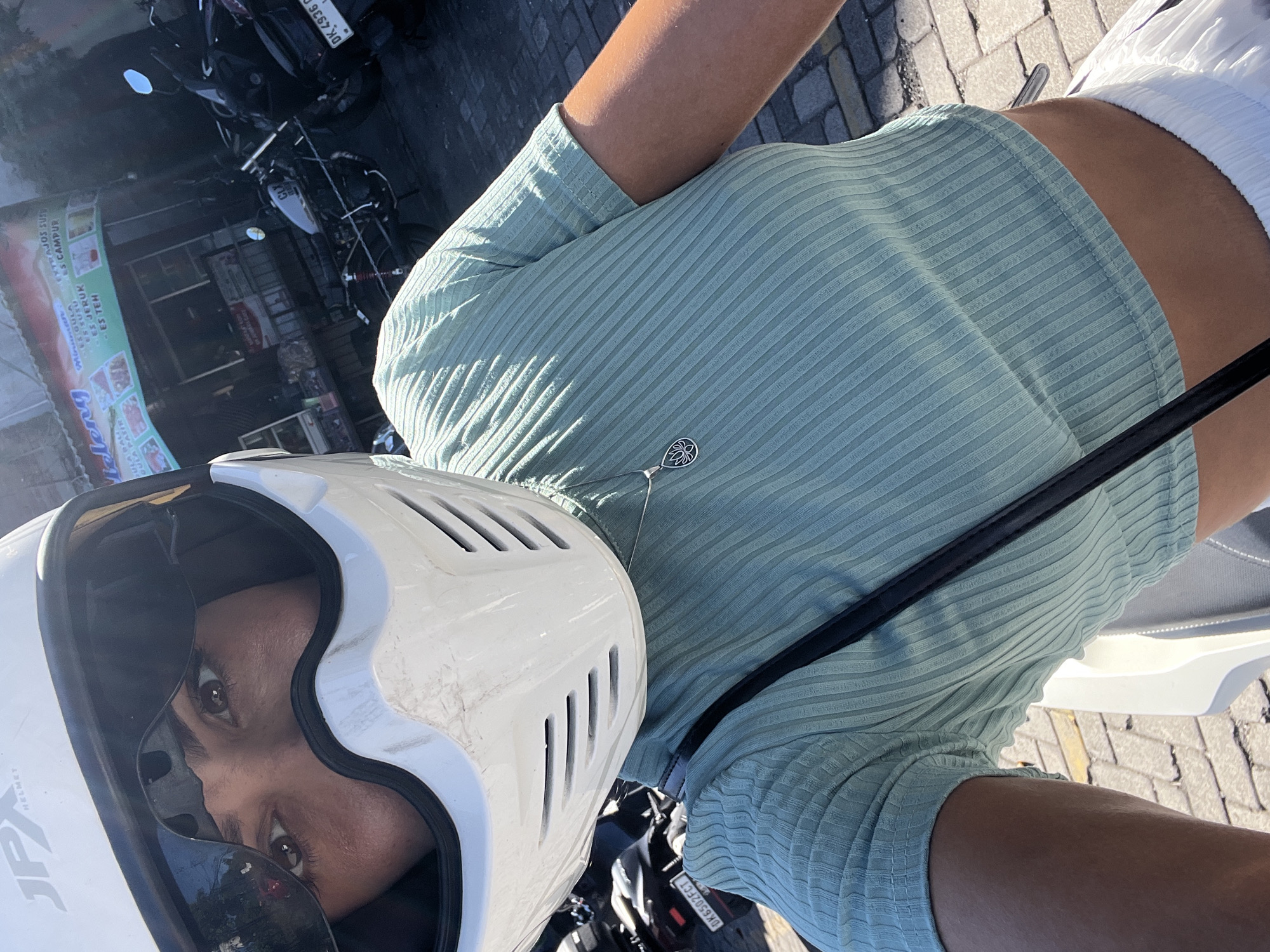 A woman wearing a white helmet and a light green ribbed top is taking a selfie on a motorcycle in Kuta Selatan, Indonesia. Visible on the left is a sign that mentions several food items including "GULA PASI," "ES GULA," "ES SUSU," "ES JERUK," "ES TEH," "EXTRAJOS SUSU," and "ES CAMPUR." Several motorcycles are parked in the background. The license plate "DK 4936" is visible on one of the motorcycles in the background, and the license plate "DK 6502FCT" is partially visible on the motorcycle the woman is on. The road is paved with interlocking bricks. The logo of "JP HELMET" can be seen on the helmet.