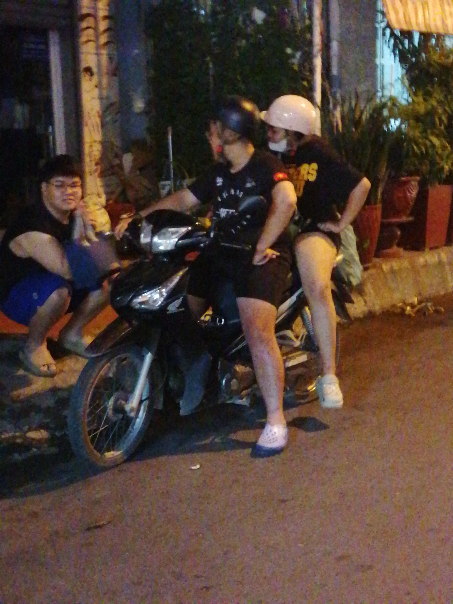 Three individuals are posing near a parked motorcycle on a city street at night. The individual on the left is a male wearing glasses, a black t-shirt, and blue shorts, sitting on an orange mat. The two individuals on the motorcycle are both wearing helmets. The person in the back is wearing a white helmet and a black t-shirt with yellow lettering. The person in the front, also wearing a black t-shirt, is partially obscured. The motorcycle is black and positioned in the center of the frame. The background shows a building with vertical beams and foliage. The ground is paved with asphalt. The scene appears to be in an urban area of Ho Chi Minh City, Vietnam, based on the presence of motorcycles and typical street architecture.
