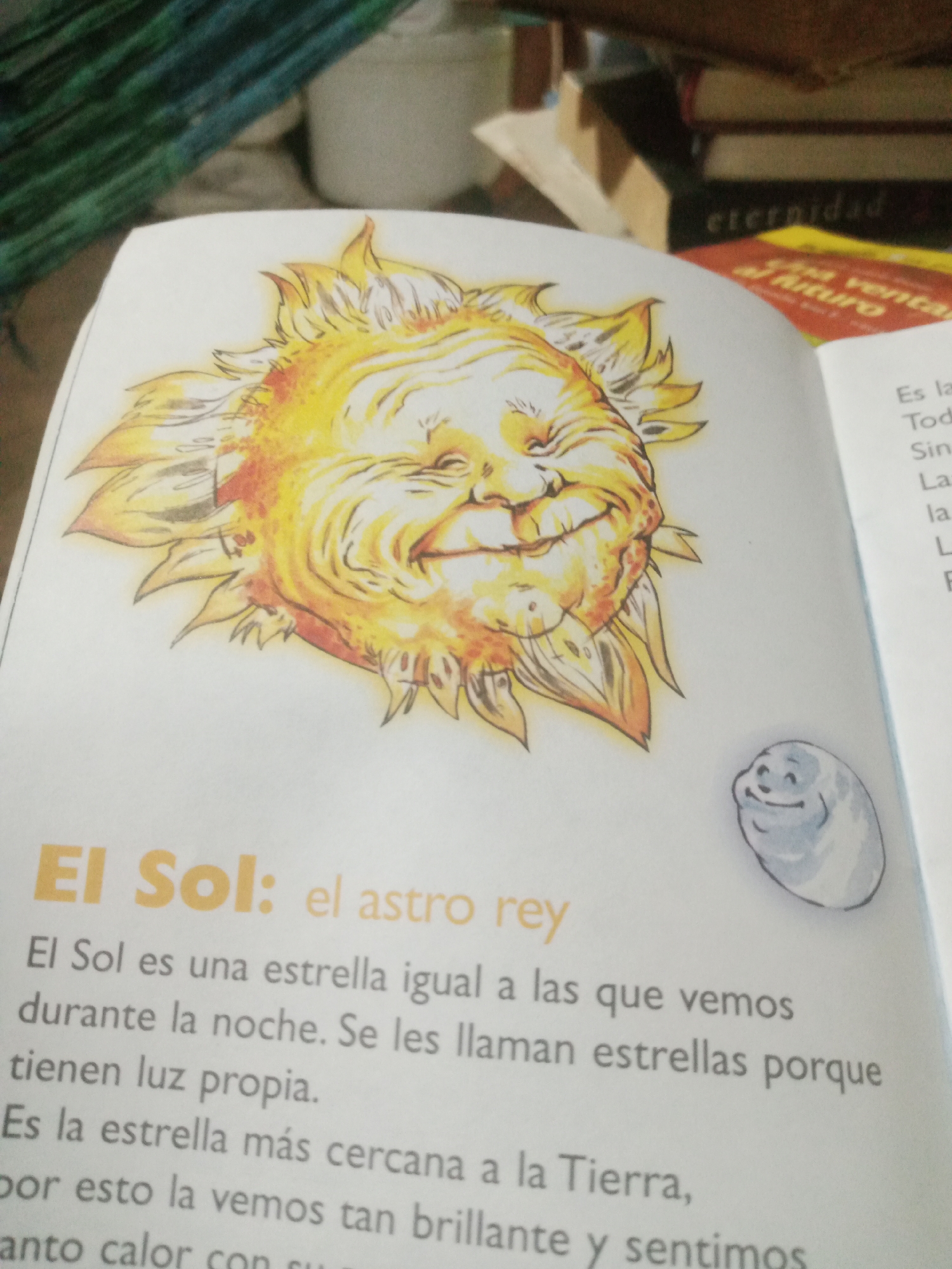 This is a close-up, eye-level photograph of an open book displaying an illustration of the sun and text. The illustration depicts a whimsical, anthropomorphic sun with a joyful, wrinkled face and fiery rays. To the lower right of the sun illustration, a small, round, blue, smiling moon-like character is drawn. The text, written in Spanish, is centered on the page and discusses the sun. The book is positioned on a surface that appears to be a wooden table, and in the background, out-of-focus stacks of books and some fabric are visible. The lighting is warm and slightly diffused, suggesting indoor lighting or a gently lit environment. There is no visible indication of the city of Santa Ana, Venezuela.