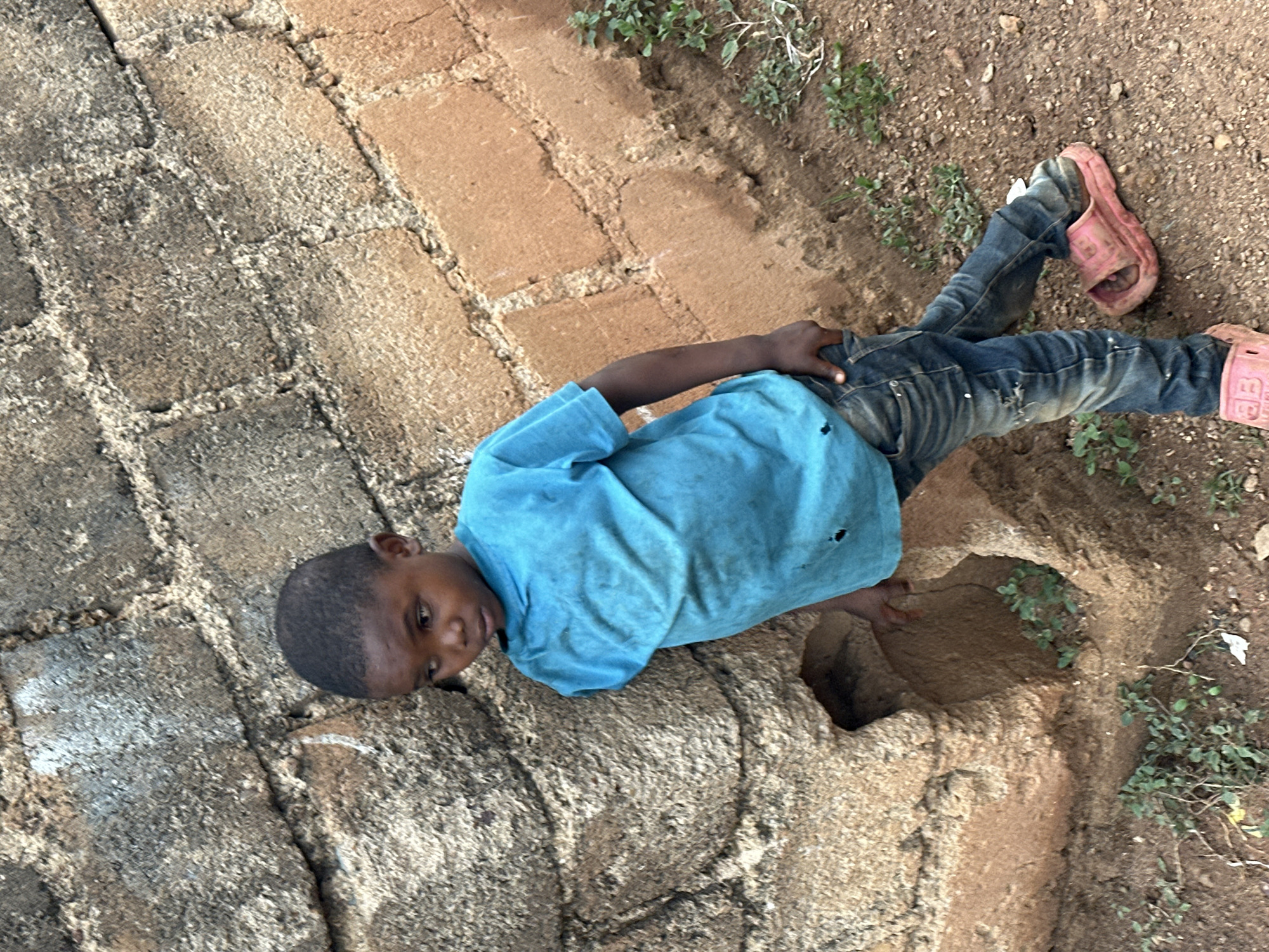 A young Black child, appearing to be a boy, stands leaning against a rough, textured wall composed of large, irregular stones and reddish-brown bricks. The child is wearing a faded blue t-shirt with small holes and distressed denim jeans. The child has one hand in their back pocket and is wearing pink slip-on sandals. The ground is covered in dirt, small stones, and sparse green vegetation. In the background, the wall continues, with a noticeable indentation or hole in the stone section. The overall scene suggests an outdoor setting in an area with simple construction. The location is identified as Gura Top, Nigeria.