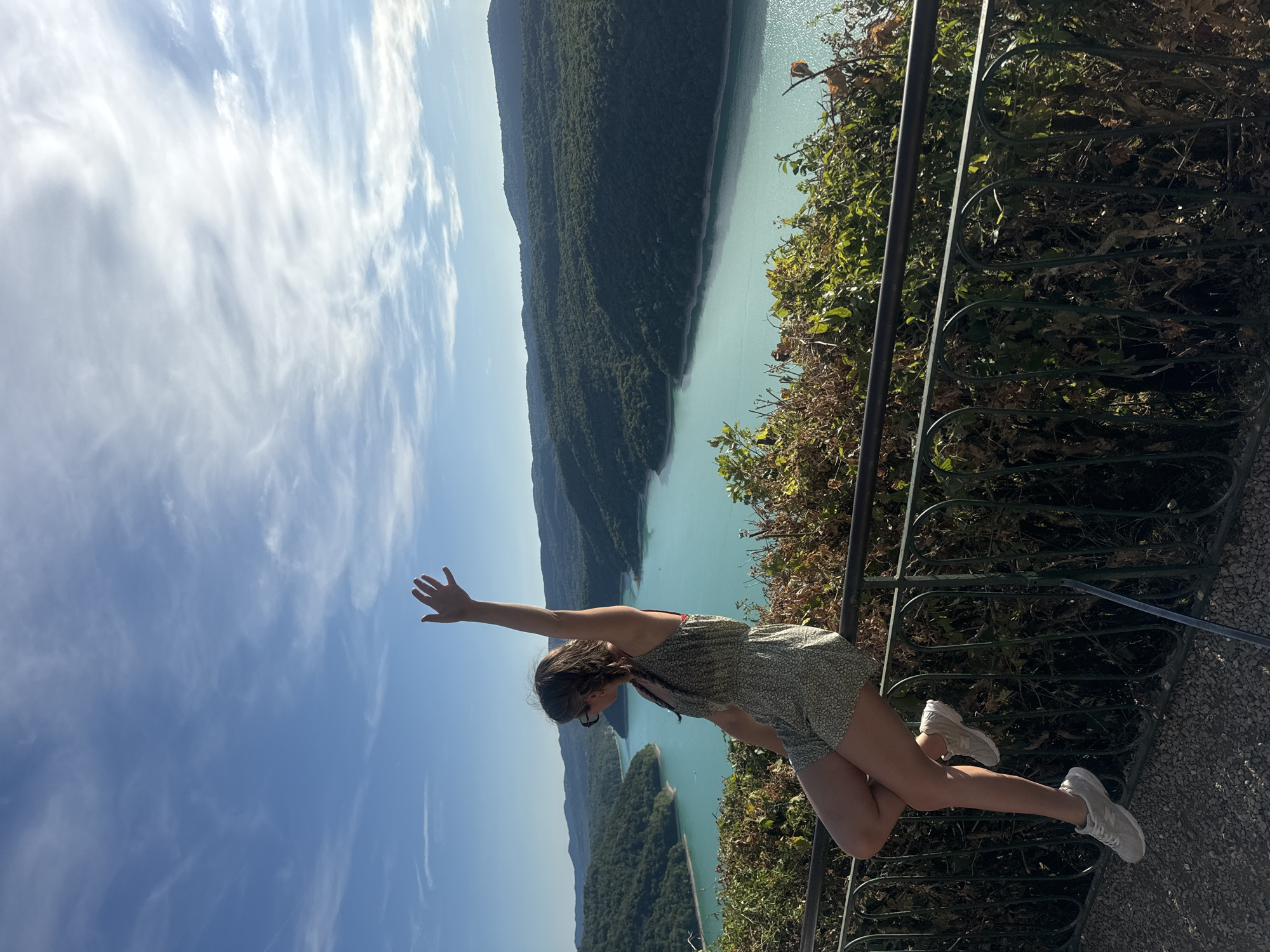 In Moirans-en-Montagne, France, a young woman is captured in a full-body, eye-level shot, perched on a black metal railing. She is facing left, with her right arm raised in a waving gesture. The woman wears a patterned romper and white sneakers. Behind her, a vast expanse of turquoise water, likely a lake or reservoir, is nestled between lush green, tree-covered hills under a bright blue sky with scattered white clouds. The scene is framed by the railing in the foreground, with dense green foliage growing behind and over it.