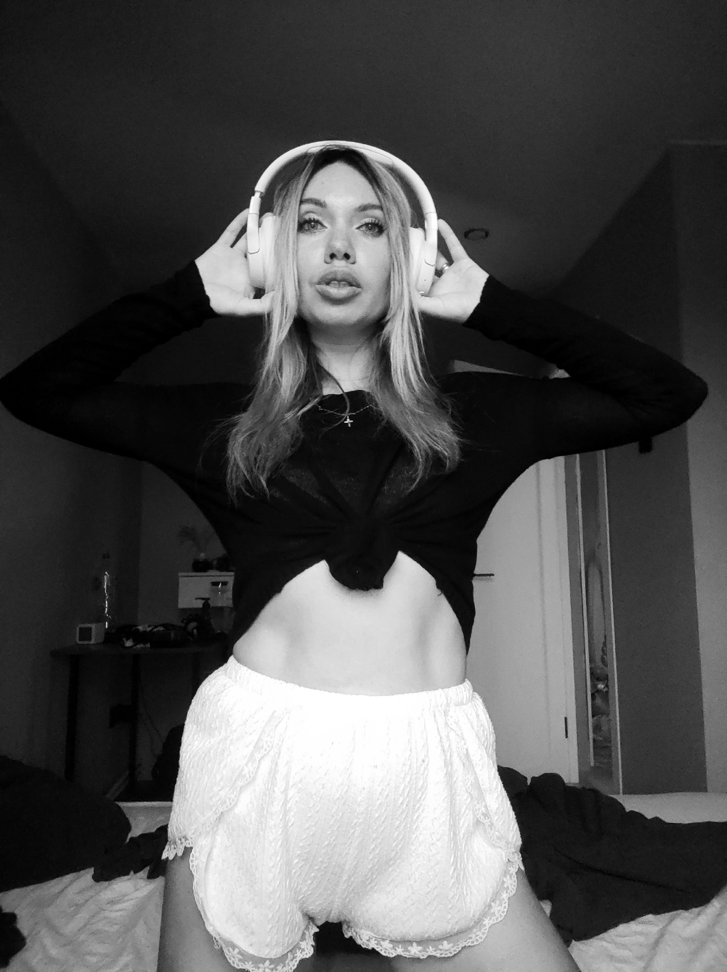 A young woman is kneeling, wearing a black long-sleeved shirt tied at the navel and white lace shorts. She has long, light hair and is wearing white headphones, with her hands holding them to her ears. The image is in black and white. In the background, there's a cluttered desk, a bed with dark blankets, and a doorway. There is no evidence to suggest this image was taken in Votkinsk, Russia.