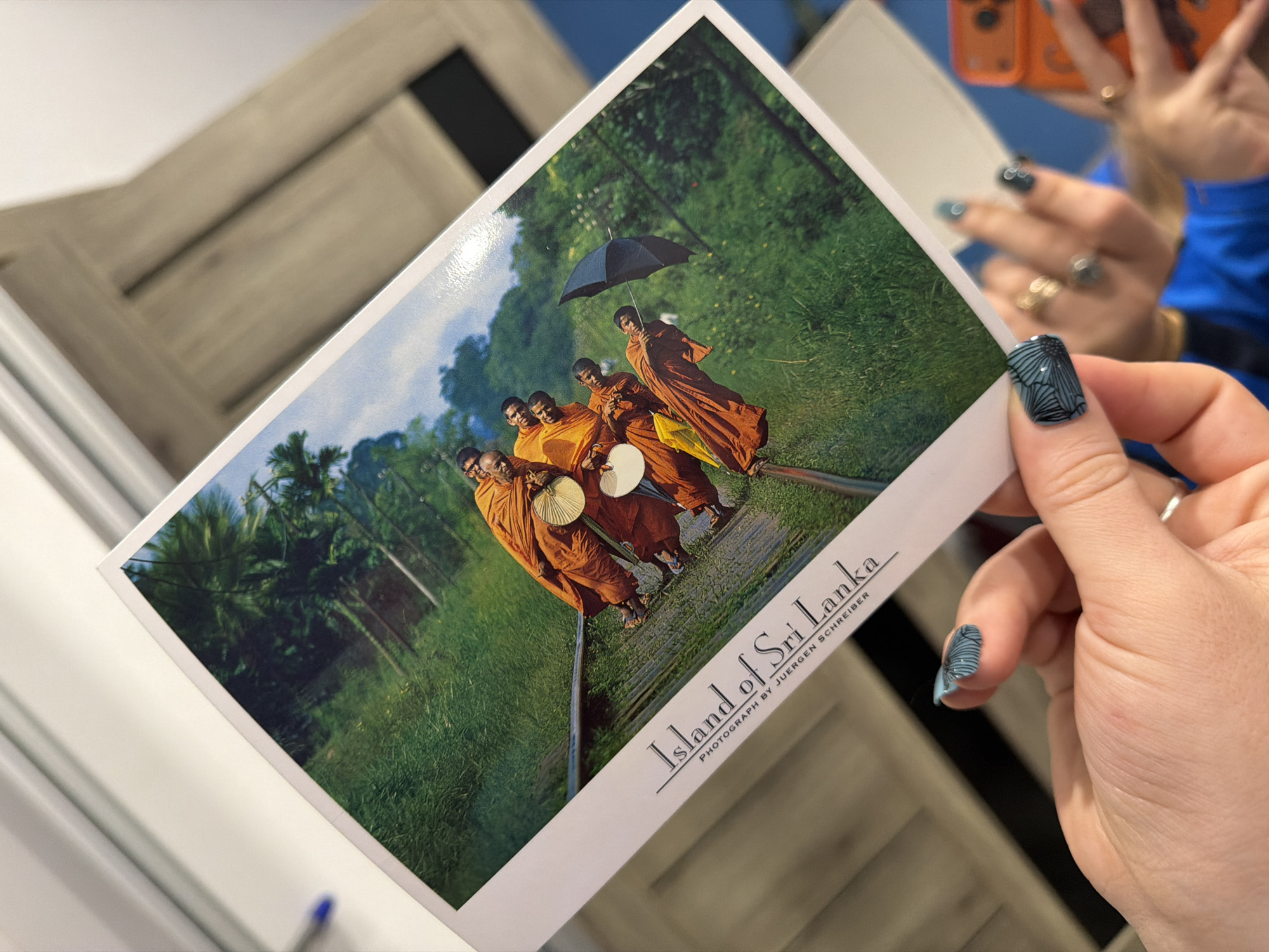 A hand holds a postcard displaying an image of five monks in orange robes walking along railroad tracks in a lush, green landscape. One monk holds a black umbrella, and another carries a white fan. The postcard text reads "Island of Sri Lanka, photograph by Juergen Schreiber". The background is blurry but appears to show a mirrored surface reflecting an orange phone and a person in a blue shirt. The location in the city of Большая Шильна, Russia is not discernible from this single image.