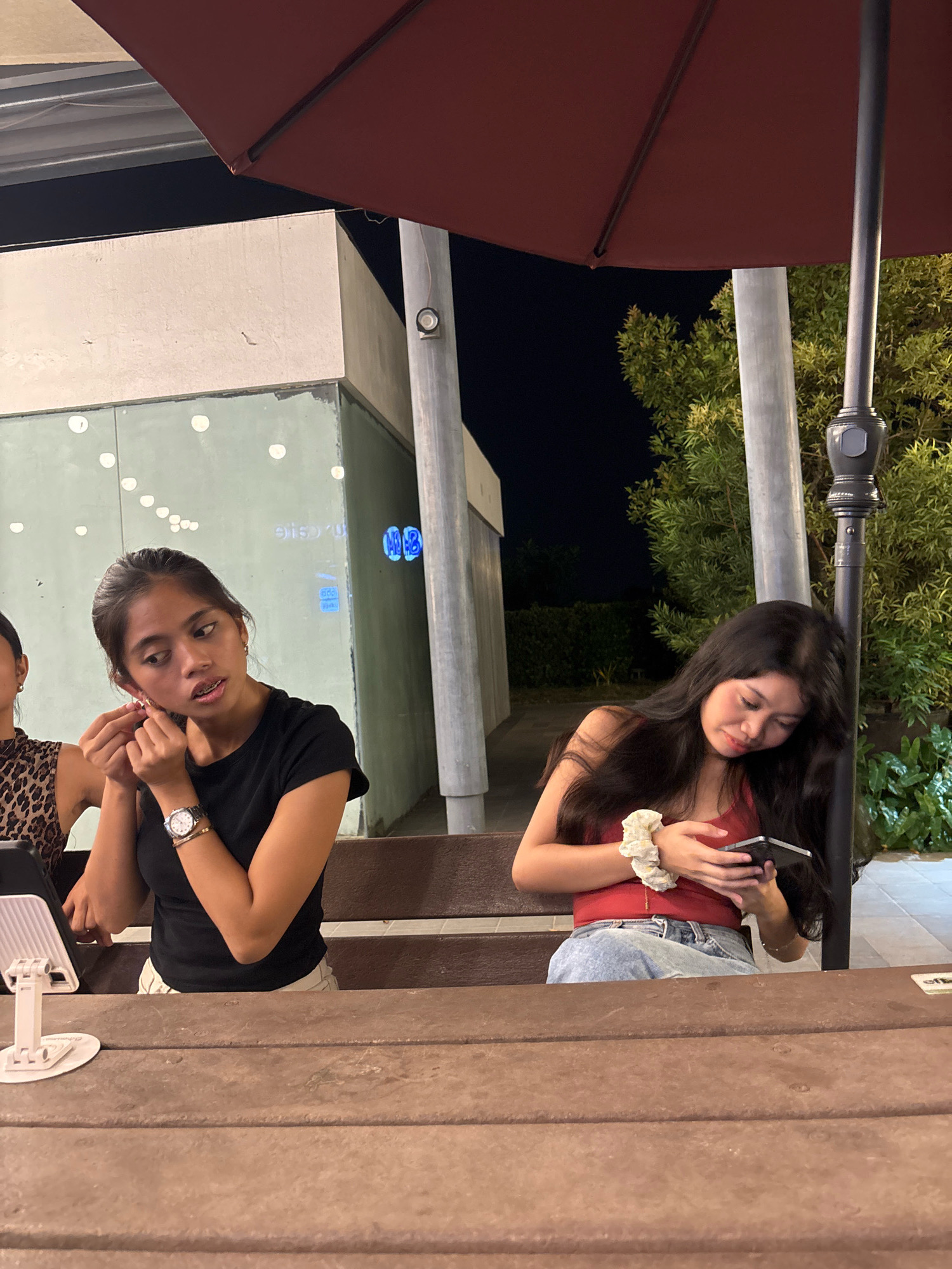 The image shows two young women sitting at a wooden picnic table at an outdoor dining establishment, likely a restaurant or cafe, in Davao City, Philippines. The woman on the left, wearing a black t-shirt, is putting on her earring. She has braces and is wearing a watch. The woman on the right, wearing a red tank top and blue jeans, is looking at her phone. She has a scrunchie on her wrist. Behind them, there is a building with a light-colored facade and large glass windows reflecting some lights, and lush green foliage. A dark red umbrella provides shade over the seating area, and a metal pole with a decorative top is visible to the right. The overall setting suggests a casual evening dining experience.