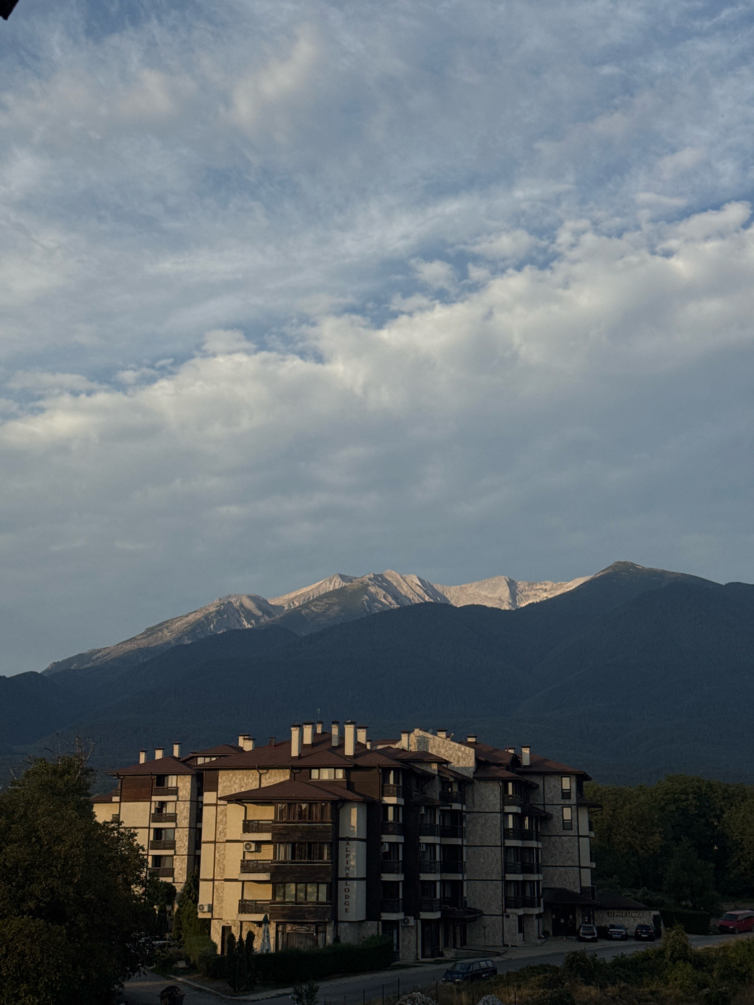 The image captures a view of the "Alpine Lodge" building in Bansko, Bulgaria, with Pirin Mountains in the background. The building is a multi-story structure with a light-colored facade, brown tiled roofs, and numerous balconies. The Pirin Mountains are dark, forested slopes leading up to rugged, rocky peaks that are partially illuminated by sunlight. The sky above is filled with a mix of scattered white clouds and darker, more overcast patches. In front of the lodge, a road is visible with a few cars parked and driving. A dense green forest borders the right side of the image, and some trees are present to the left of the lodge. The overall scene suggests an early morning or late afternoon setting due to the lighting conditions.
