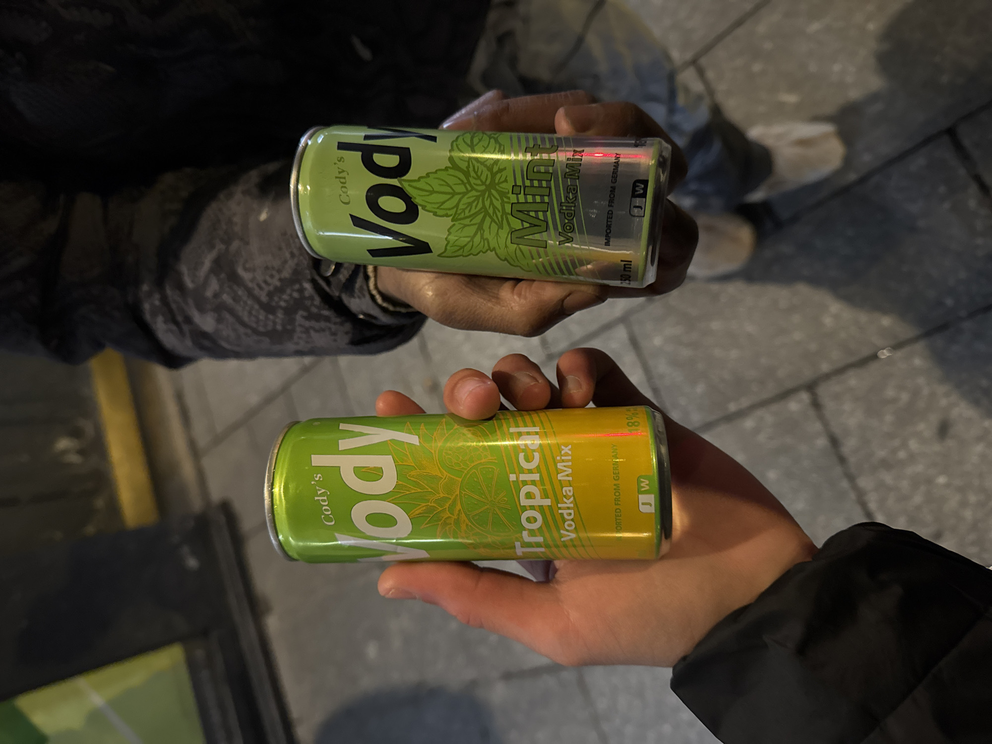 Two individuals are depicted holding canned alcoholic beverages outdoors on a paved surface in Paris, France, under artificial illumination. A hand with a light skin tone, adorned with a dark sleeve, is centrally positioned, holding a 250 ml 'Cody's Vody Tropical Vodka Mix' can. This can features green and yellow coloring with fruit graphics, states '18%' alcohol content, and is marked 'IMPORTED FROM GERMANY'. To the right, a hand with a darker skin tone holds a 250 ml 'Cody's Vody Mint Vodka Mix' can, which is green and silver with mint leaf graphics, also indicating '18%' alcohol and 'IMPORTED FROM GERMANY'. The individual holding the mint can is partially visible in the background, wearing dark clothing and light footwear. The ground consists of grey rectangular paving stones. A blurry green and yellow object is visible in the upper left background.