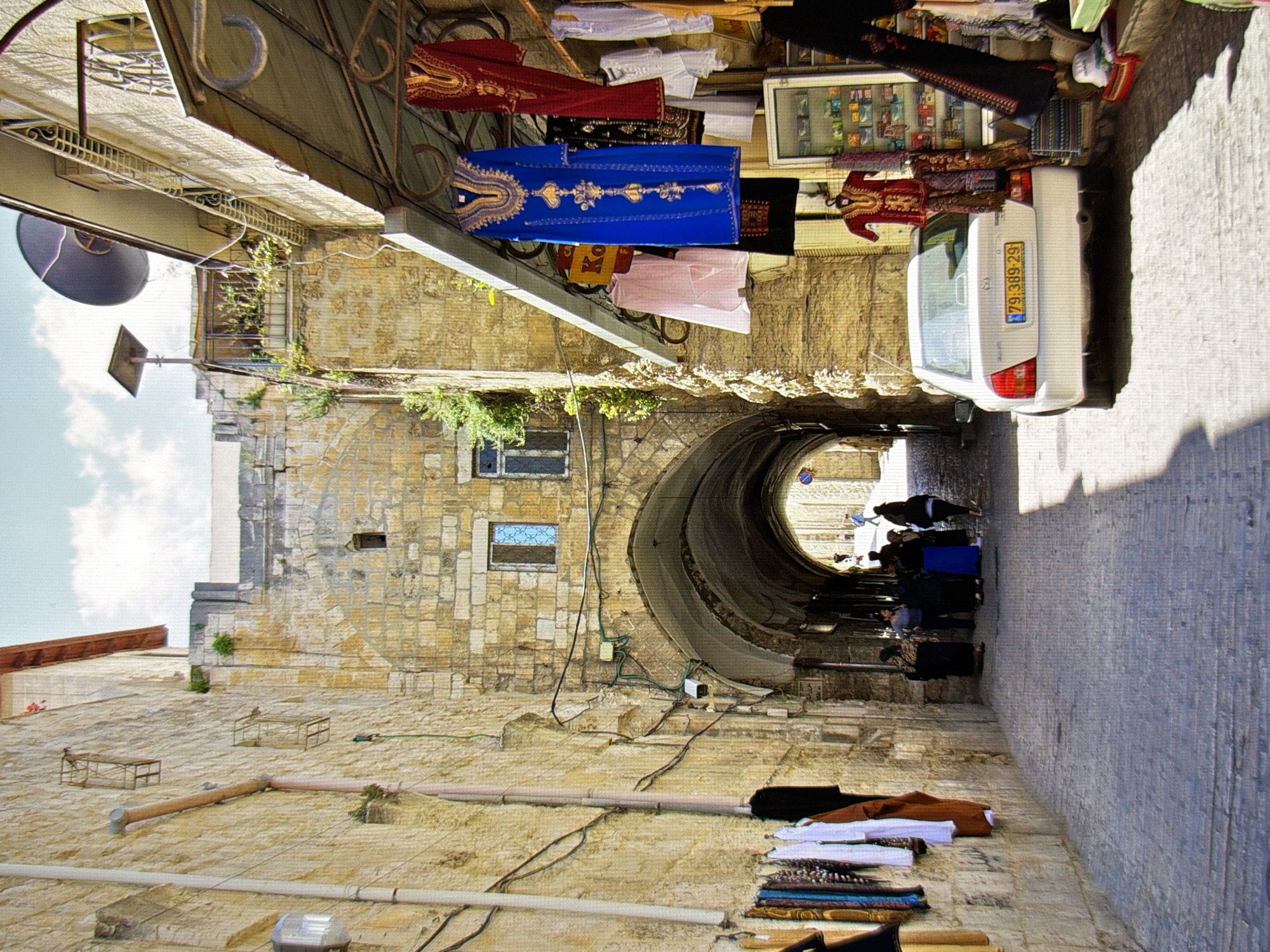 The image depicts a narrow, stone-paved alleyway in a historic city, likely in the Middle East, not Kyiv, Ukraine. The alley is lined with old stone buildings with arched doorways and windows. On the left side, clothing is hanging on display, attached to the side of a building. On the right side, there is a shop with clothing and merchandise displayed outside, including several colorful dresses and other textiles. A white car with a license plate "79 389 29" is parked in the alley. Several people are visible, some walking through the alley and others standing near the shops. The sunlight creates shadows on the ground, indicating it is daytime. The architecture suggests a location with significant historical and cultural heritage.