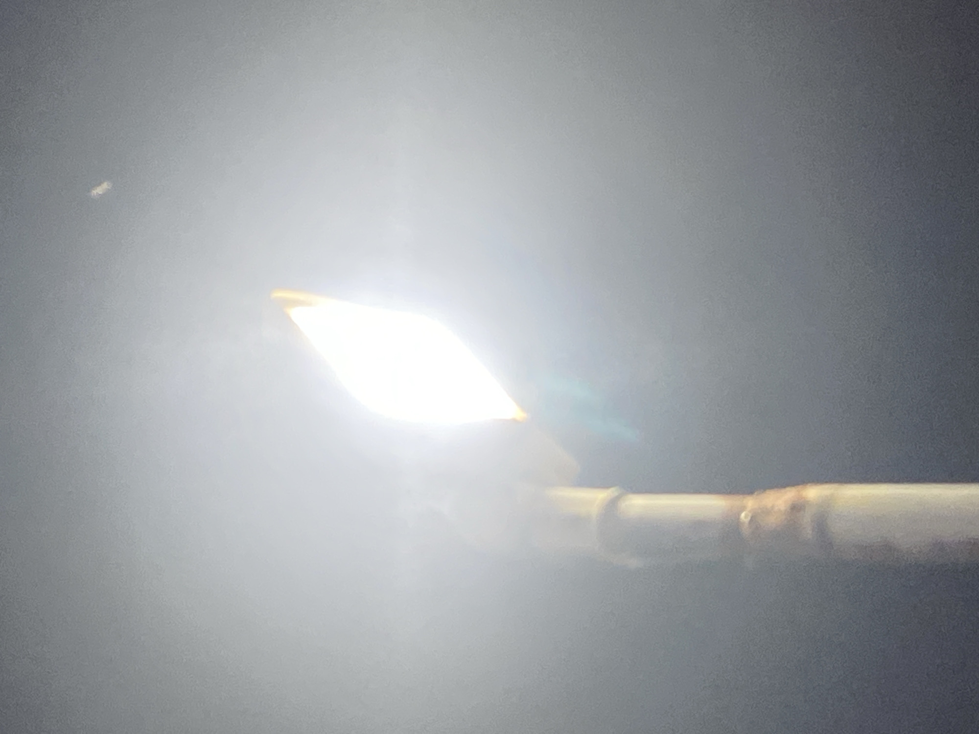 The image depicts a brightly illuminated outdoor light fixture, likely a streetlamp, emitting an intense white-yellow light against a dark, ambient background. The light source itself is overexposed, producing significant lens flare and a bright halo effect that fades into a dark grey-black environment, characteristic of nighttime conditions. A vertical, cylindrical pole, appearing light-colored, supports the light fixture. Approximately midway down the visible section of the pole, an irregular, brownish-orange band or wrapping is present, with a small, lighter circular protrusion visible on the pole's surface just above this band. In the upper right quadrant of the frame, a single, faint point of light is visible. No individuals or specific actions are depicted. The image contains no visual elements or contextual information that specifically identify its location within the city of Ikorodu, Nigeria.
