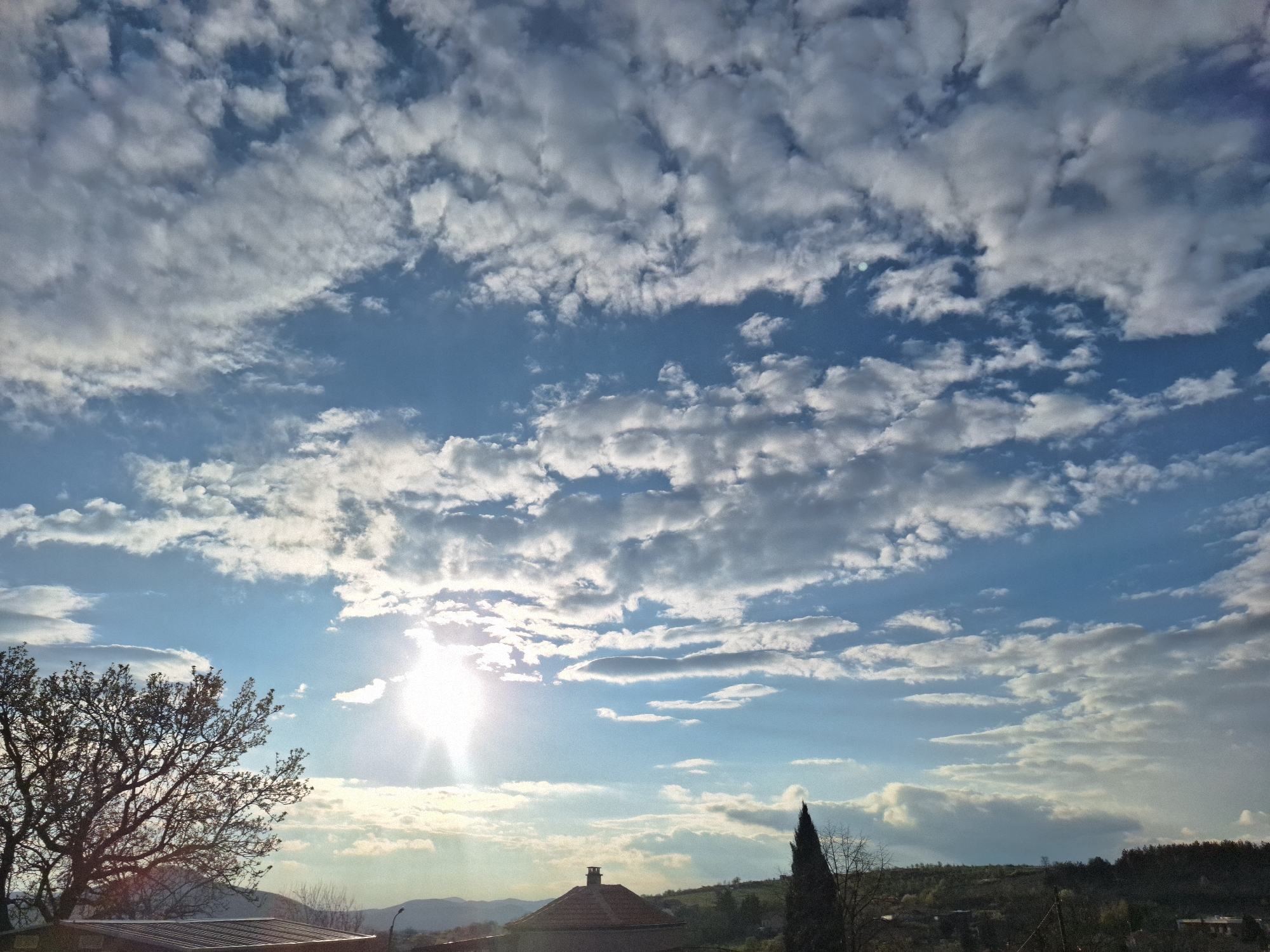 The image captures a daytime scene, likely outdoors, under a sky filled with scattered clouds and patches of blue. The sun shines brightly, creating a lens flare effect and illuminating the clouds. In the lower portion of the frame, there are elements suggestive of a cityscape or suburban area, including building rooftops and possibly some vegetation. The perspective suggests a ground-level view looking upwards.