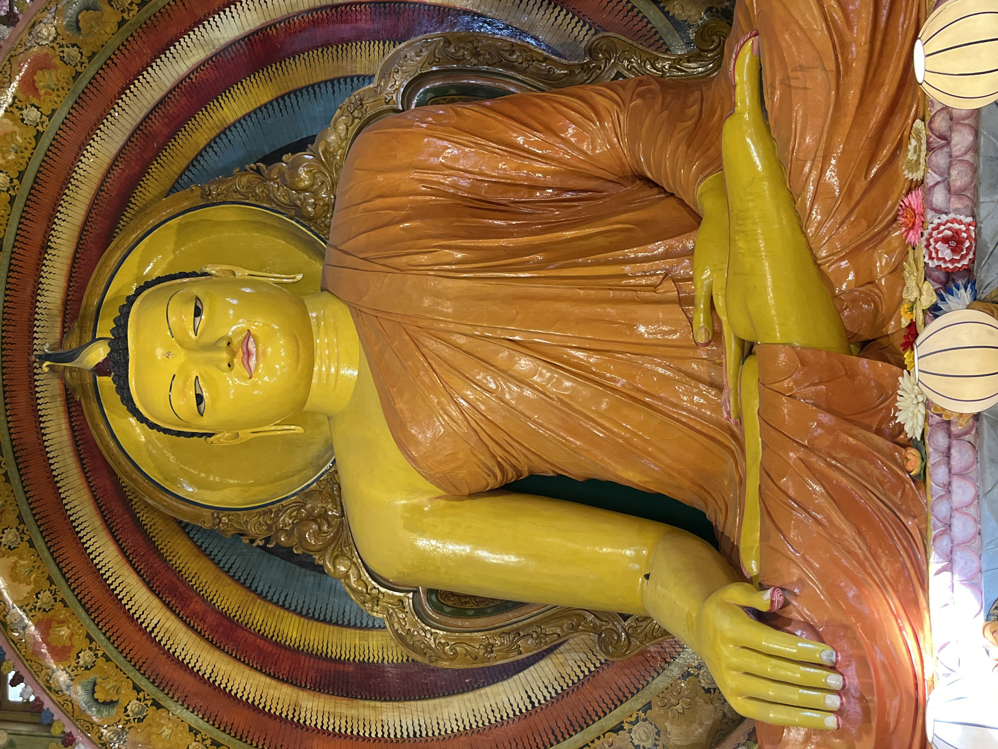 The image shows a large, yellow, seated Buddha statue in a brightly decorated shrine. The statue is adorned in orange robes and sits with its legs crossed, hands resting in its lap. The background features an elaborate, radiating halo or mandorla with multiple colorful concentric circles and decorative motifs. At the base of the statue, there are offerings of flowers and decorative lanterns. The statue is likely located in a Buddhist temple or temple complex in Colombo, Sri Lanka, given the style of religious art and the commonality of such statues in the city. The lighting suggests an indoor setting, possibly illuminated by artificial light sources and natural light filtering in.