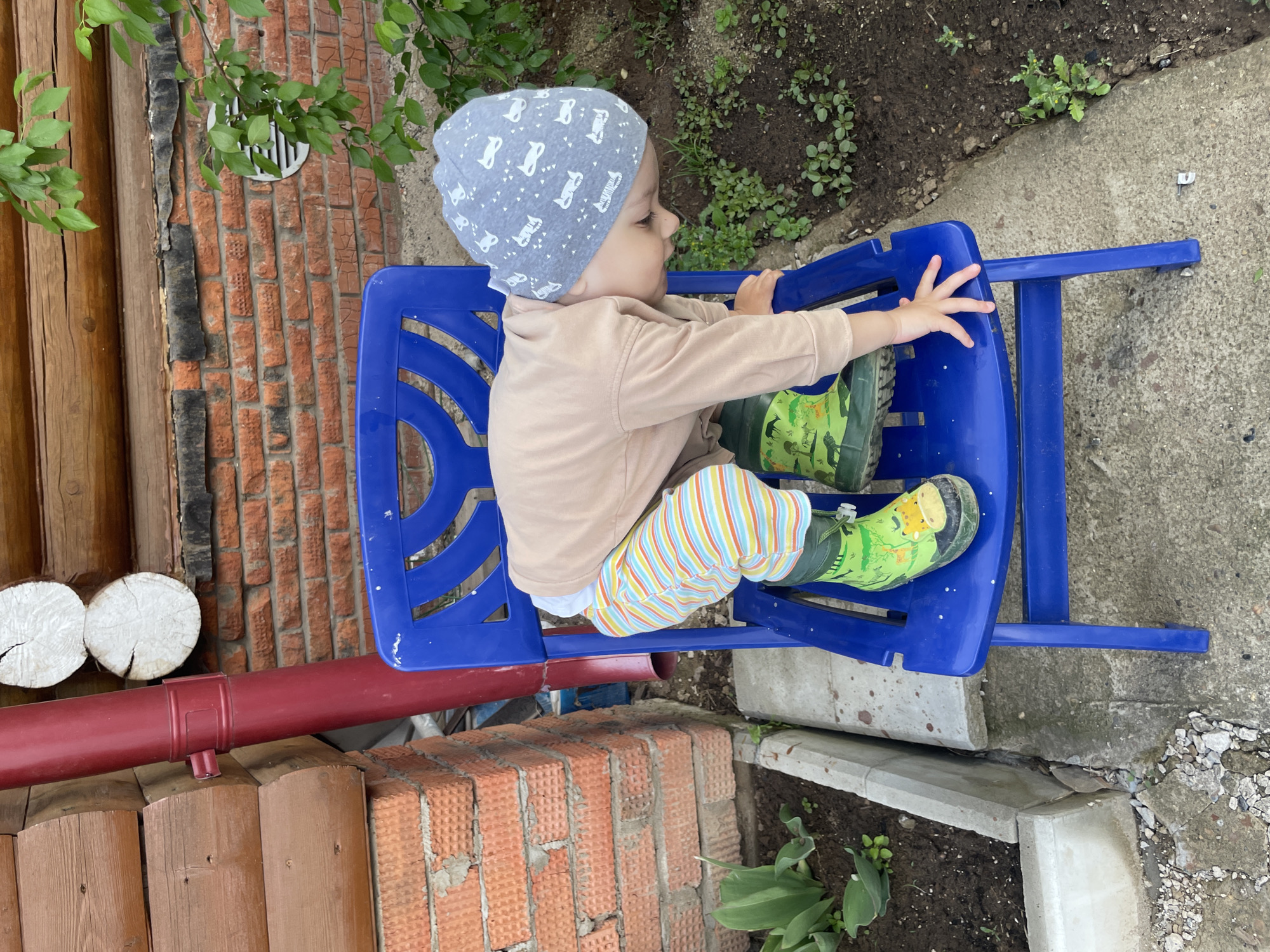 A child wearing a gray penguin-patterned hat, beige long-sleeve shirt, colorful striped pants, and green animal-print boots is seated on a blue plastic chair outdoors. The child is facing away from the camera and reaching forward with their right hand. The setting includes elements of a house, with red brick walls, a dark red pipe running horizontally, and cut logs visible in the background. A concrete path and dirt area with green plants are also present. This image was likely taken in Zav'yalovo, Russia.