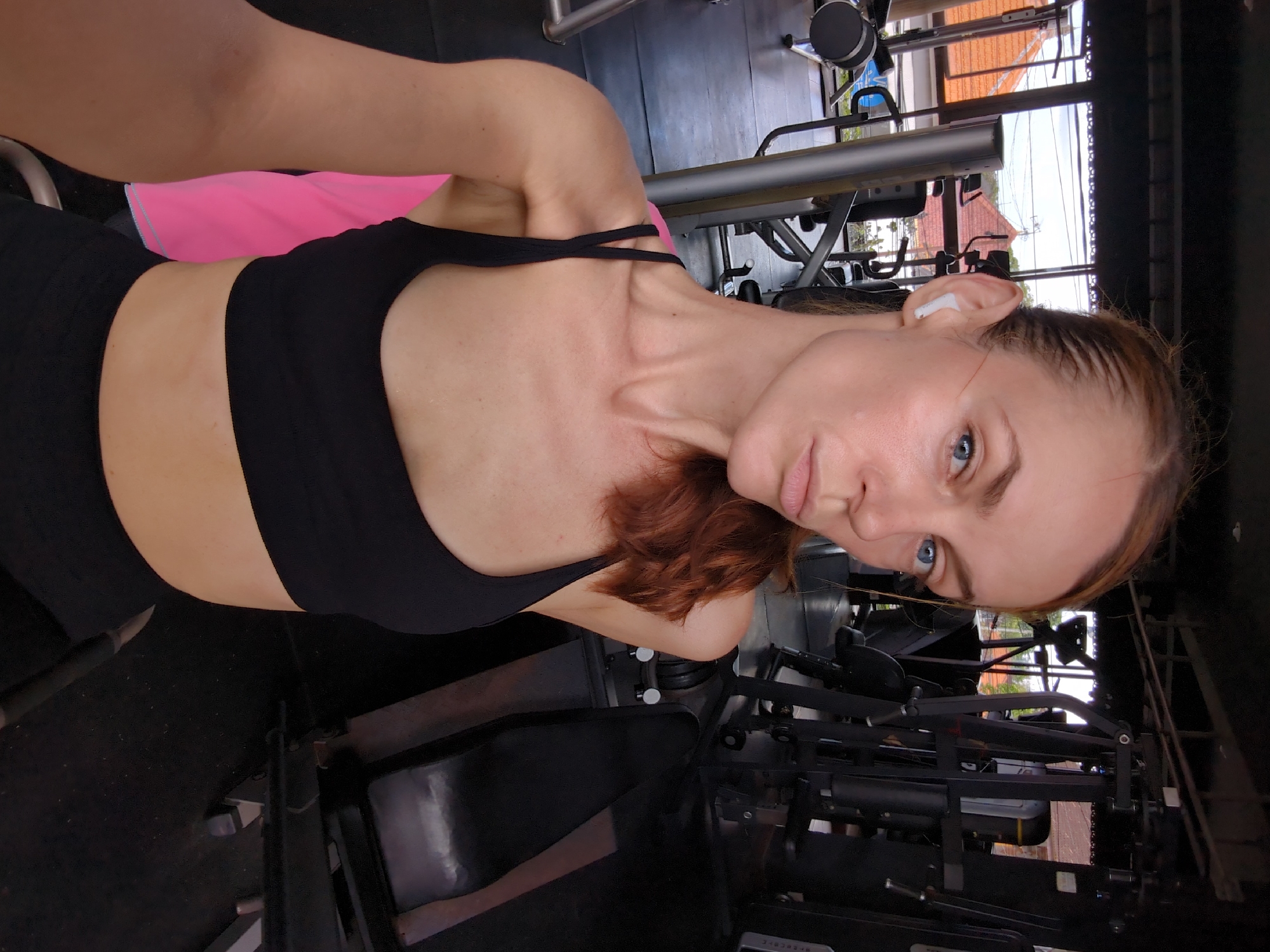 A woman is at a gym, wearing a black sports bra and black shorts. She has a pink towel draped around her neck. She is wearing a wireless earbud in her right ear. The gym is located in Mengwi, Indonesia. Weight machines are visible. Outside the windows, roofs and trees are visible.