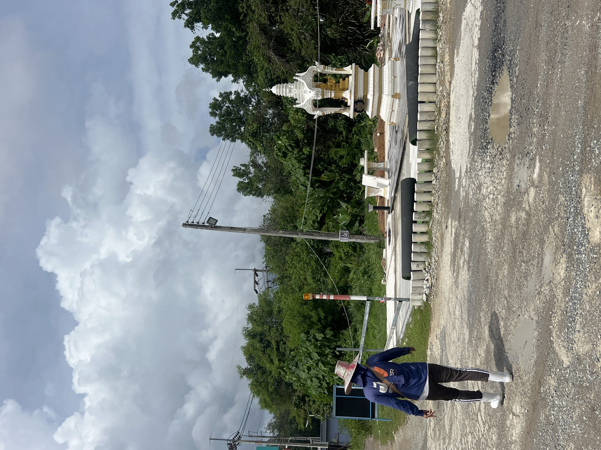 The image depicts a person walking away from the camera on a gravel road in Pa Khlok, Thailand. The person is wearing a blue long-sleeved shirt, dark pants with white stripes, a wide-brimmed hat, and white boots. They are carrying a brown bag slung over their shoulder. To the right of the person, there is a small decorative shrine with white and gold accents, which appears to be in a newly constructed or renovated area. The area around the shrine is paved with stone tiles and bordered by a low fence made of concrete posts and dark cylindrical objects. Beyond this area, lush green trees form a dense background. In the foreground, the gravel road has several puddles of water, indicating recent rain. Utility poles and power lines are visible against a cloudy sky. A blue sign board is present to the left of the frame. The overall scene suggests an outdoor location that may be a public space or a construction site undergoing development.