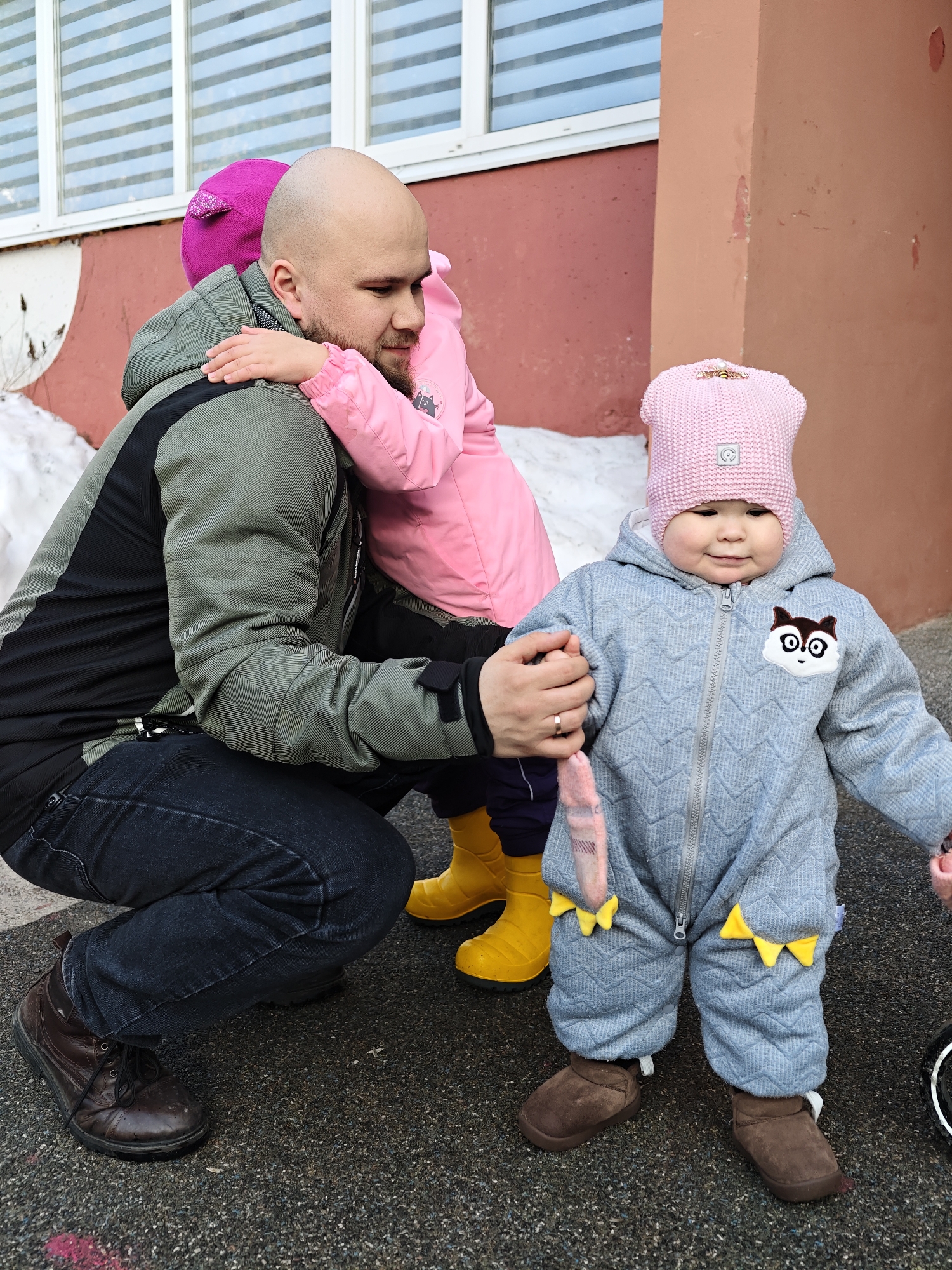 The scene depicts three individuals outdoors in the city of Chaykovsky, Russia, against a building facade. The building features a reddish-brown lower section and a lighter upper section with a window displaying horizontal blinds. A patch of snow is visible on the ground to the far left. The ground surface is dark gray asphalt.
An adult male, bald with a beard, occupies the center-left. He wears an olive green and black patterned jacket, dark blue jeans, and brown lace-up shoes, with a gold-colored ring on his left ring finger. He is crouched on his left knee.
An older child, partially obscured behind the adult male, wears a magenta knit hat, a pink jacket, and yellow boots. This child embraces the adult male, with arms wrapped around his upper back.
A younger child, identified as a toddler, stands foreground-right, facing the viewer with a slight smile. This child wears a light pink knit hat with a small emblem and a golden barrette, and a light gray hooded jumpsuit with a chevron texture, a front zipper, a white and brown animal applique on the left chest, and yellow triangular accents on the lower sleeves. The child wears brown soft-soled shoes and holds a pink item in their right hand while their left hand is held by the adult male's right hand.
The lighting is bright and even, indicating an outdoor setting in cold weather conditions.