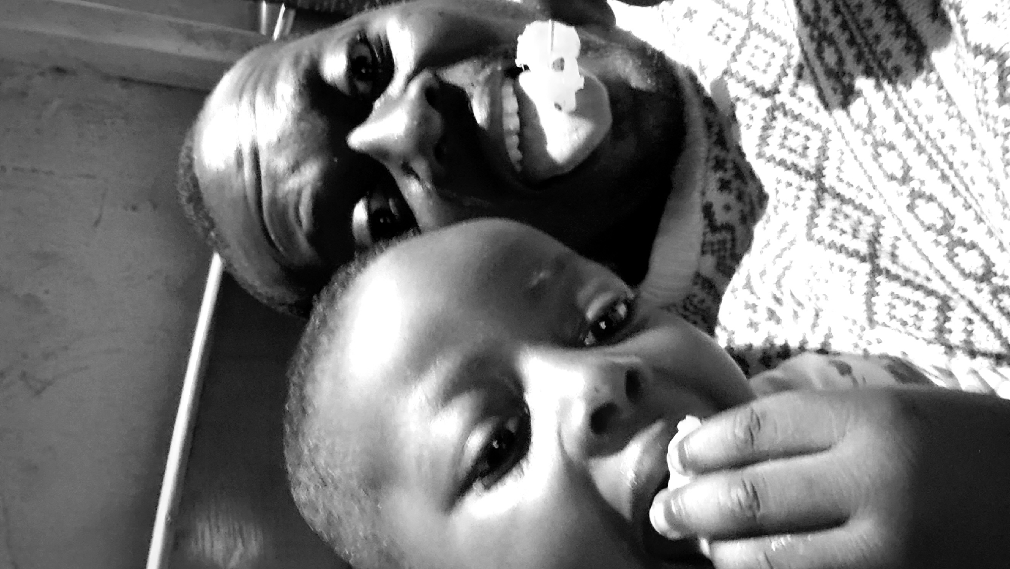 The image is a close-up, black and white, selfie-style shot featuring two individuals, an adult male and a child, in what appears to be an outdoor setting in Sabon Tasha, Nigeria. The adult male is on the right, his face taking up a significant portion of the frame, with his mouth open and his tongue extended, showing a piece of white material, possibly candy or food, on his tongue. His eyes are wide open, and his facial expression suggests a playful or surprised demeanor. The child is positioned on the left, their head closer to the camera, with their left hand near their mouth, appearing to be eating or holding something. The child's eyes are also open, and their gaze is directed towards the camera. Both individuals are wearing patterned clothing, the adult's shirt having a geometric design. The background is blurred but suggests an overhead structure, possibly a roof or awning, with some texture visible. The lighting indicates it is daytime, with strong shadows. The overall scene conveys an informal and intimate moment shared between the two individuals.