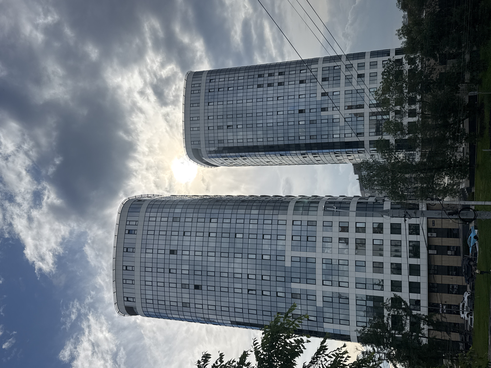 The image depicts two modern, cylindrical high-rise buildings with a facade of glass and white panels, situated in the city of Izhevsk, Russia. The sun is partially obscured by dramatic, dark clouds, creating a contrast between bright light and shadow. Trees and foliage are visible in the foreground and surrounding the buildings, suggesting an urban setting with green spaces. Electrical wires crisscross the sky, indicating the presence of urban infrastructure. Several cars are parked near the base of the buildings, and there is a glimpse of street-level retail spaces. The overall impression is one of contemporary architecture set against a dynamic, overcast sky.